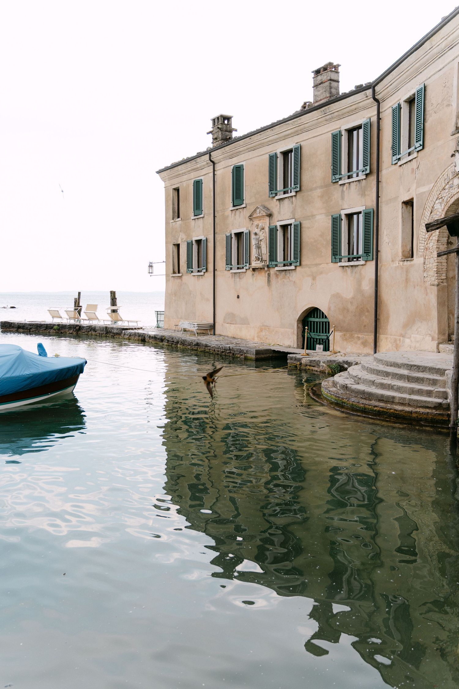 A series of historic waterfront buildings with green shutters reflect in the calm canal waters of Venice, Italy.