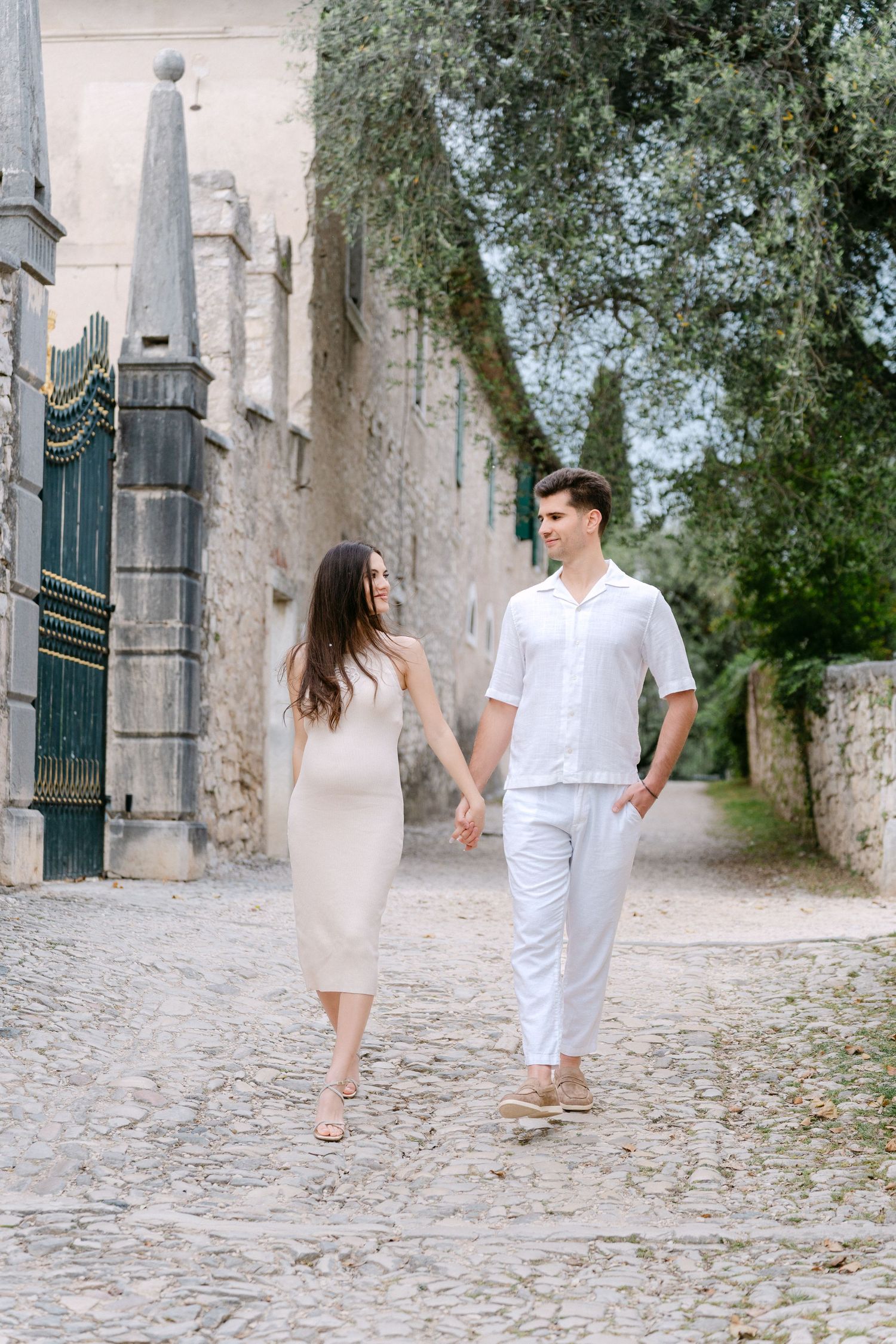 A couple in matching white outfits walks hand in hand along a cobblestone path beside an old stone wall.