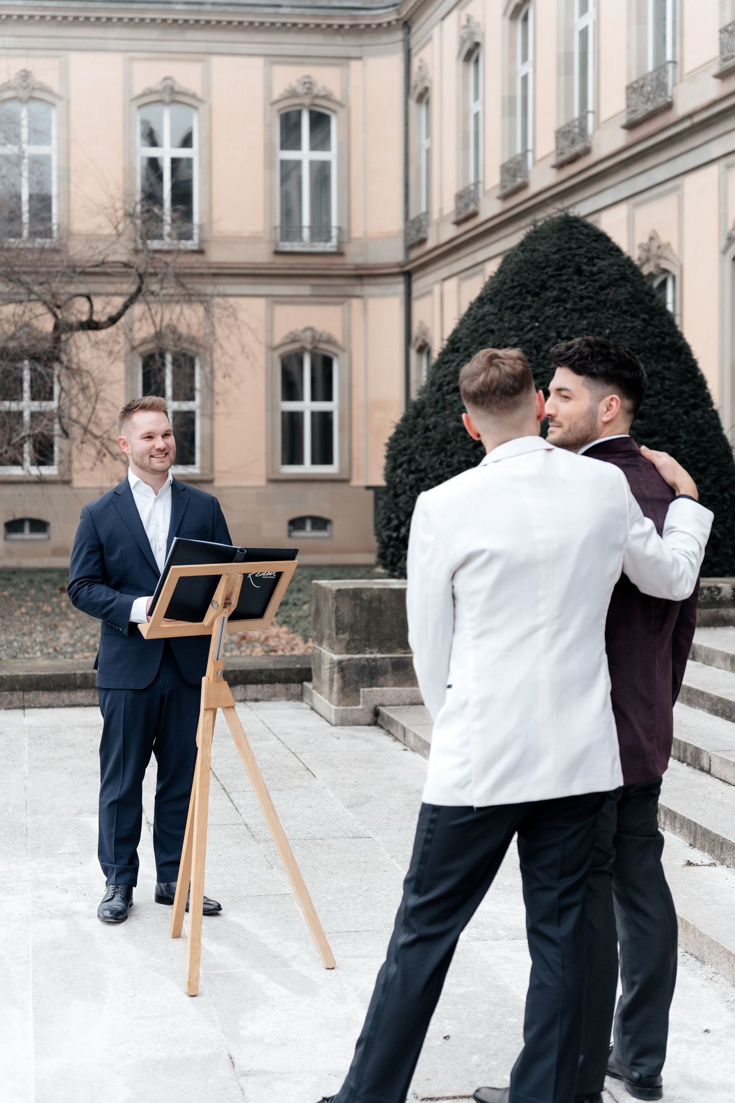 An artist sketches with a wooden easel outdoors in front of a historic European building with arched windows.
