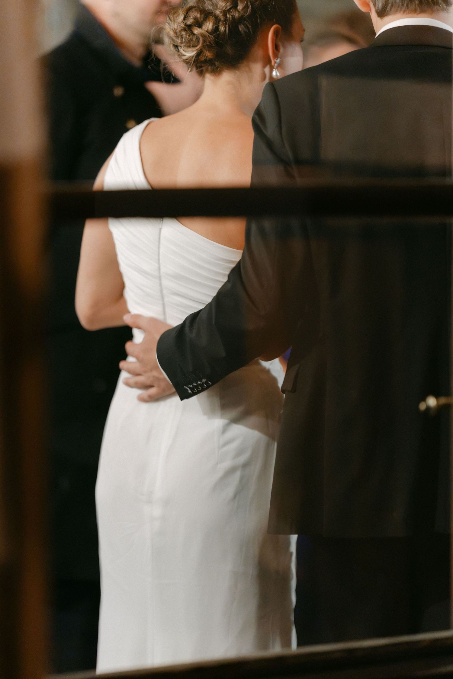 Bride in an elegant white one-shoulder gown with an updo stands at the altar as the groom in a dark suit rests his hand on her back.