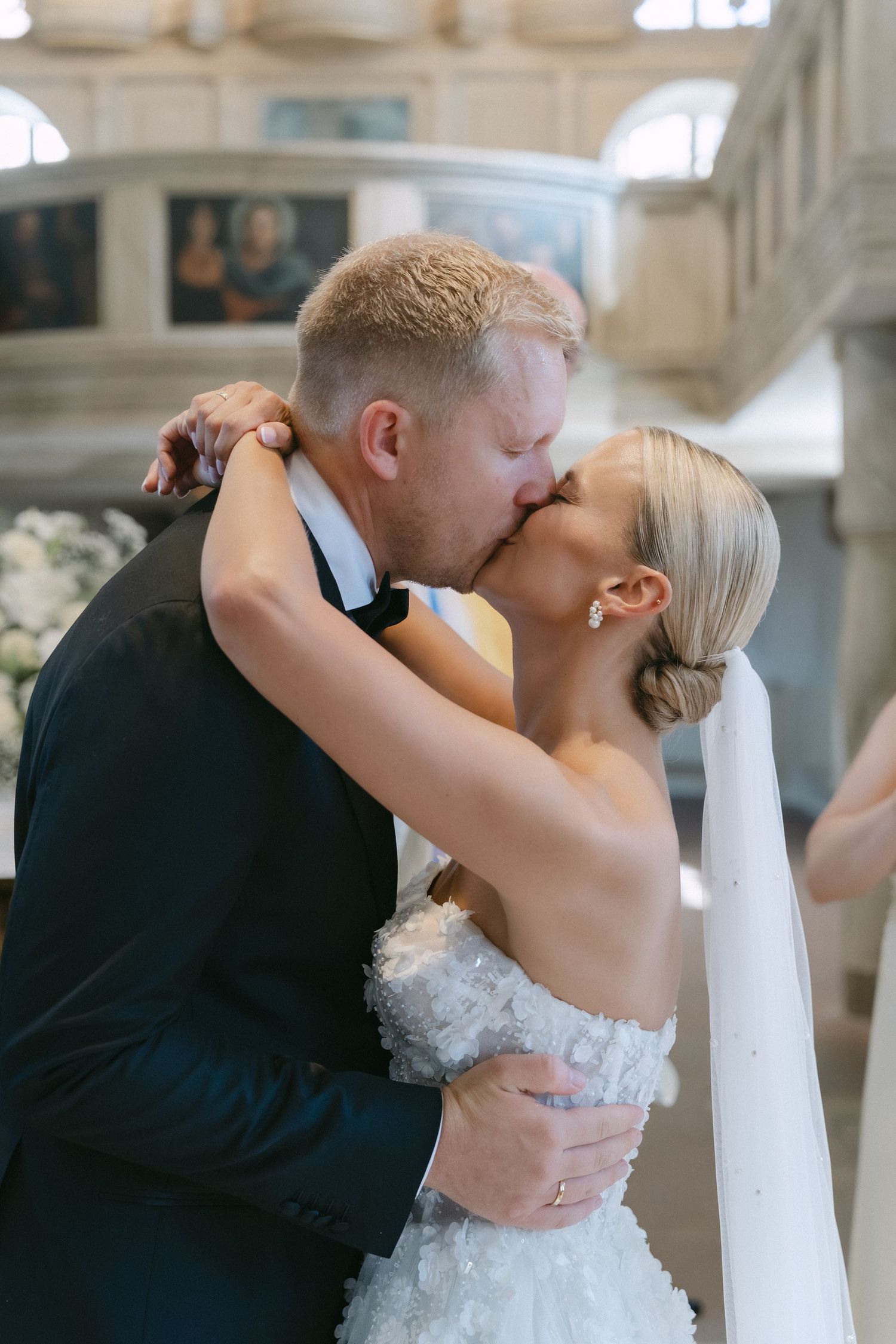 Bride in a white floral gown and veil kisses her groom in a dark suit during their wedding ceremony inside a church.