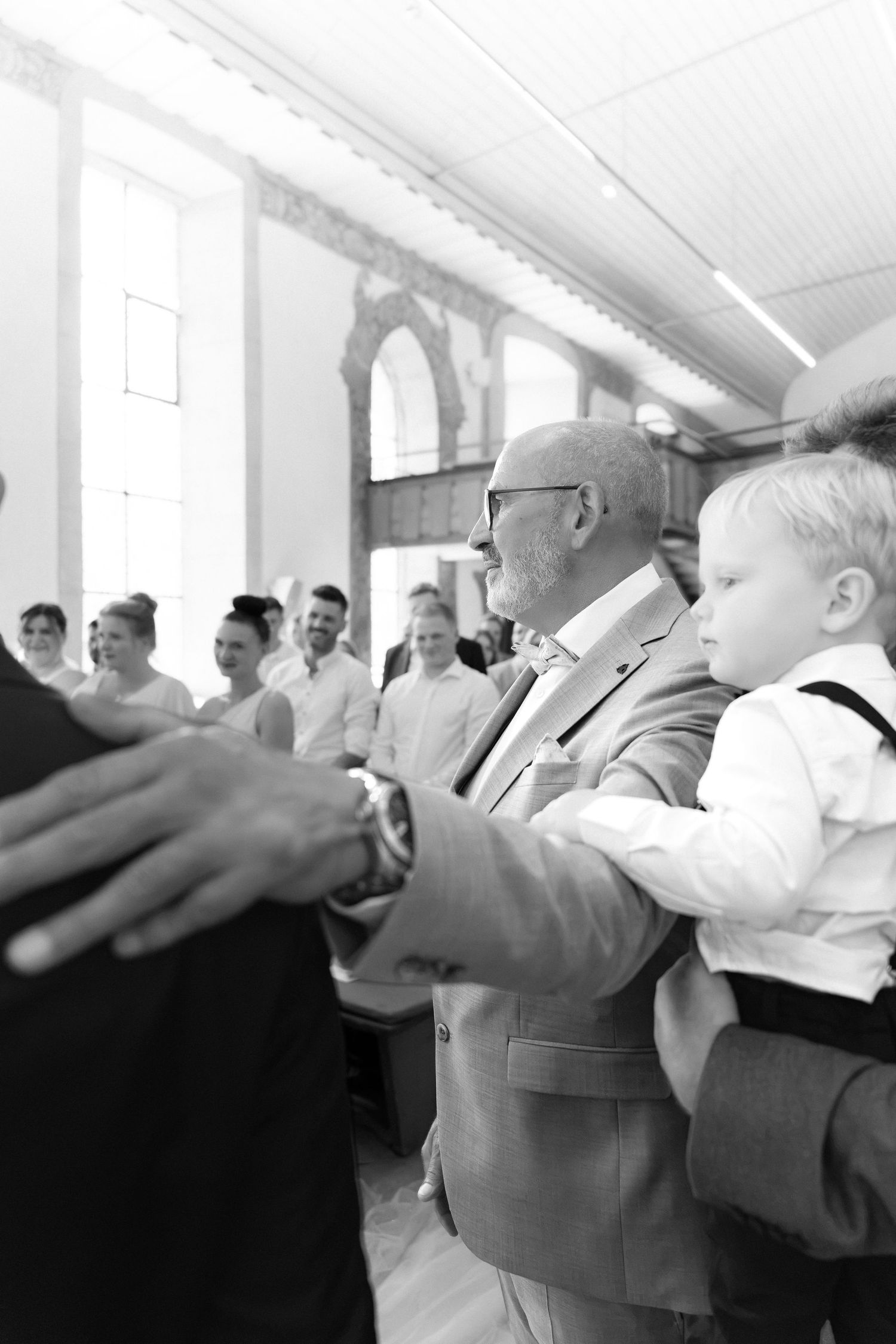 Black and white photo of a man in glasses and a blazer holding a young boy at a wedding gathering, with guests smiling behind them.