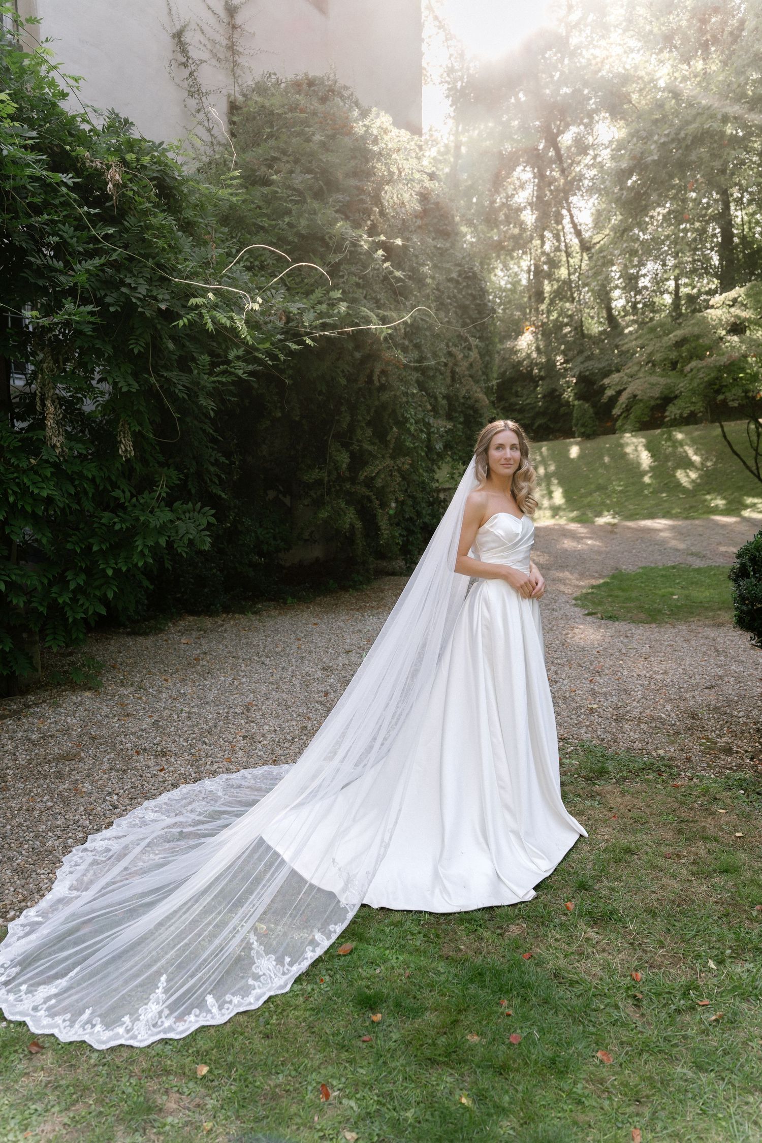 Bride in a strapless satin wedding gown with a long lace-edged cathedral veil stands on a gravel path in a sunlit green garden.