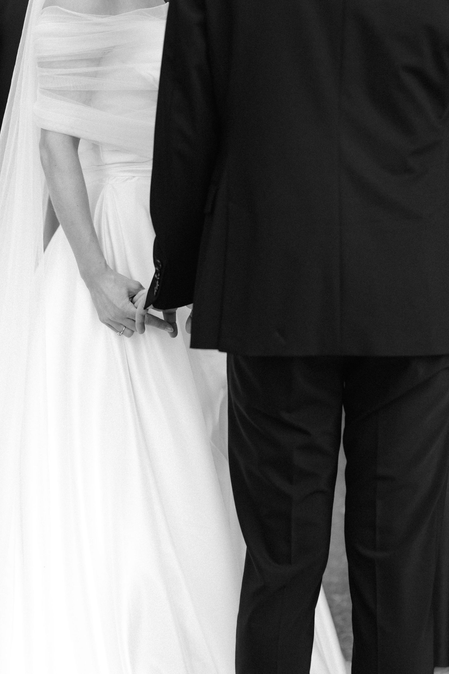 Black and white photo of a bride in a white gown and veil holding hands with a groom in a dark suit, seen from behind.