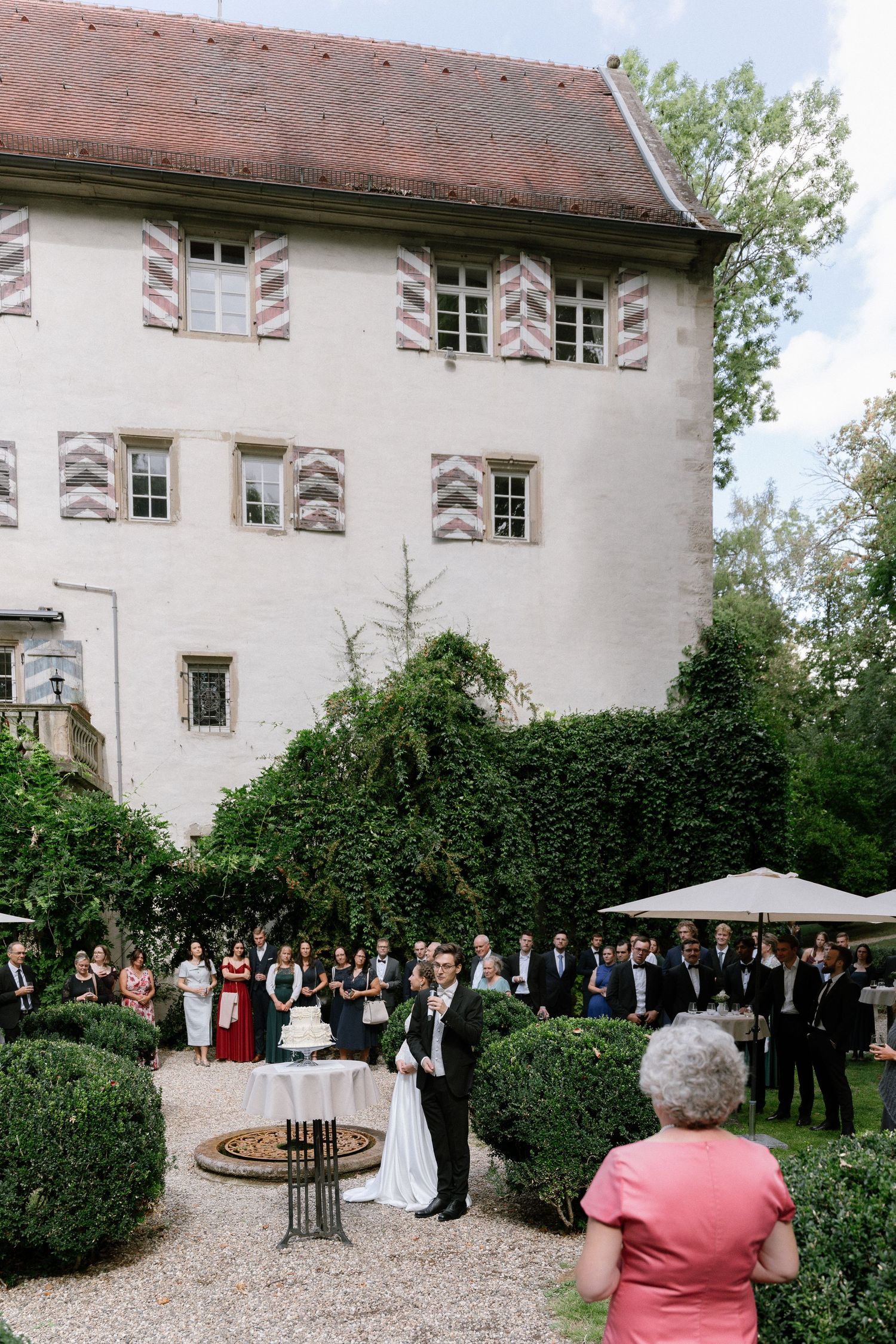 Wedding guests gather on a gravel courtyard around a couple and cake table beside a historic white castle with striped shutters.