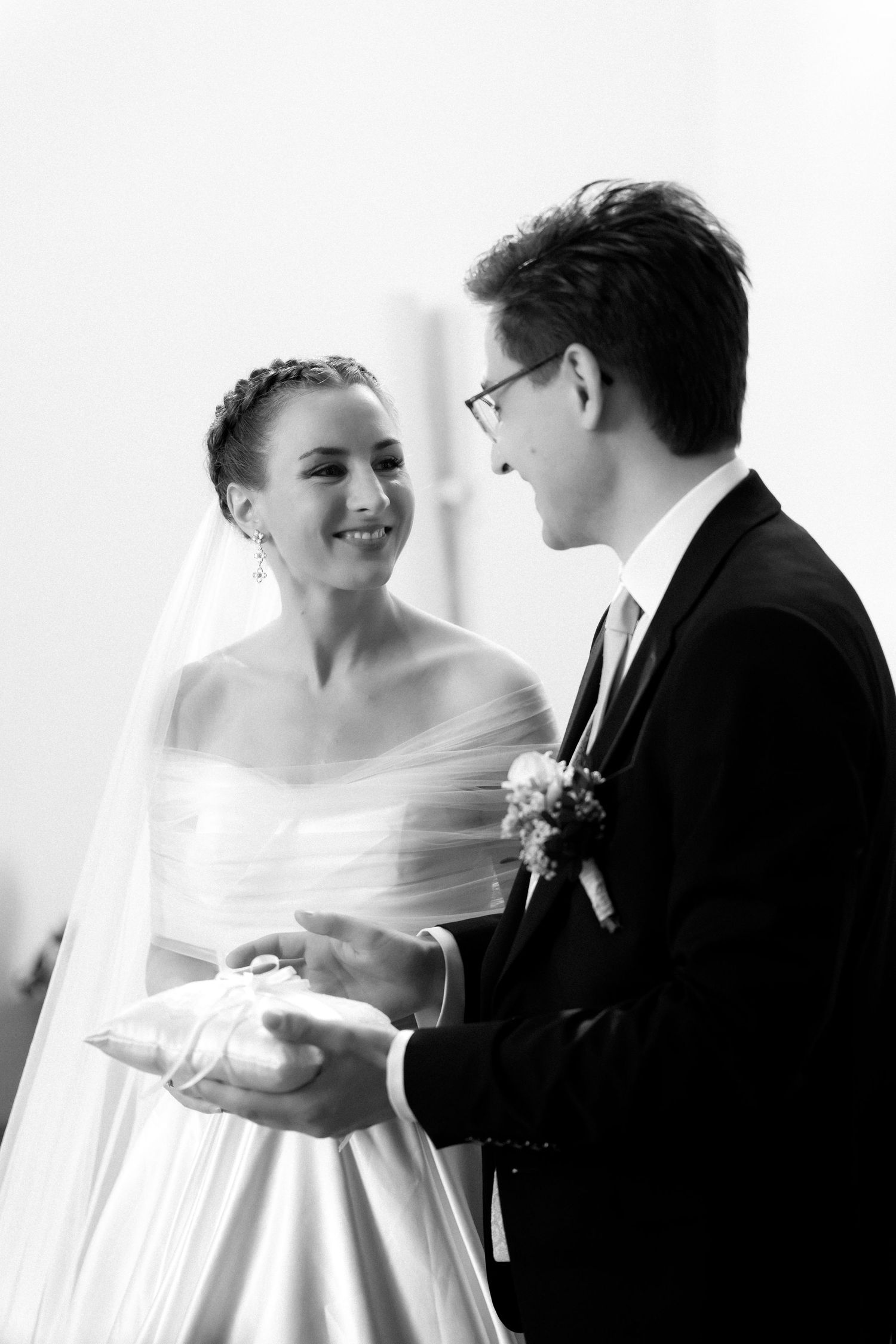 Smiling bride in a veil and strapless gown holds a ring pillow with the groom in glasses and a boutonniere during their wedding ceremony.