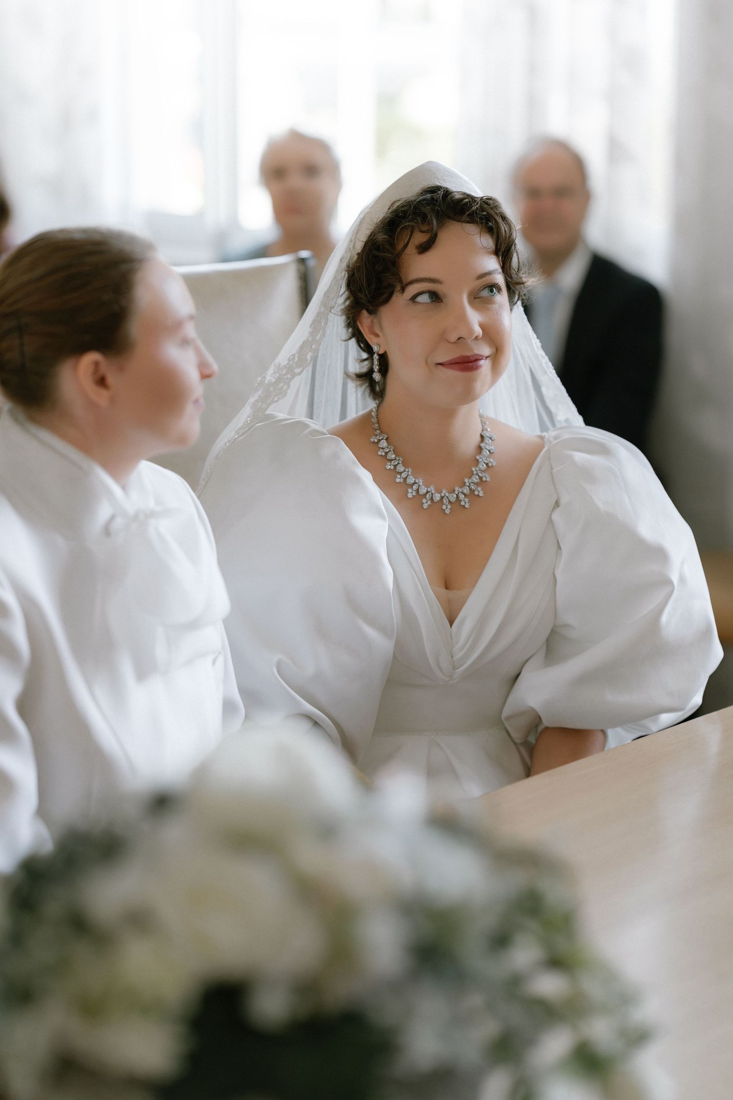 Bride in white gown with veil and diamond necklace sits at ceremony table, guests visible in background.
