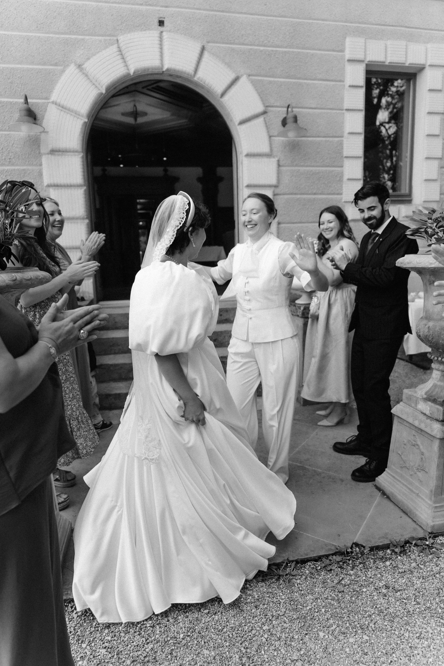 Black and white photo of a bride in a flowing gown and veil being celebrated by wedding guests outside a venue.