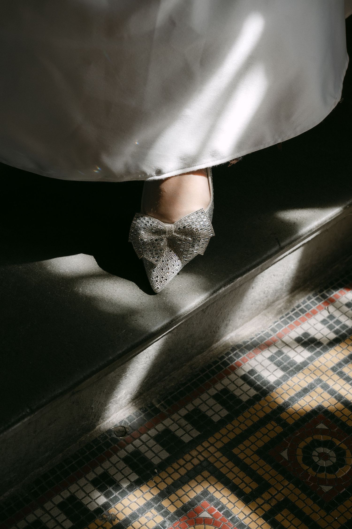 Bride wearing sparkling bow-embellished wedding flats on mosaic tile stairs beneath a white gown.