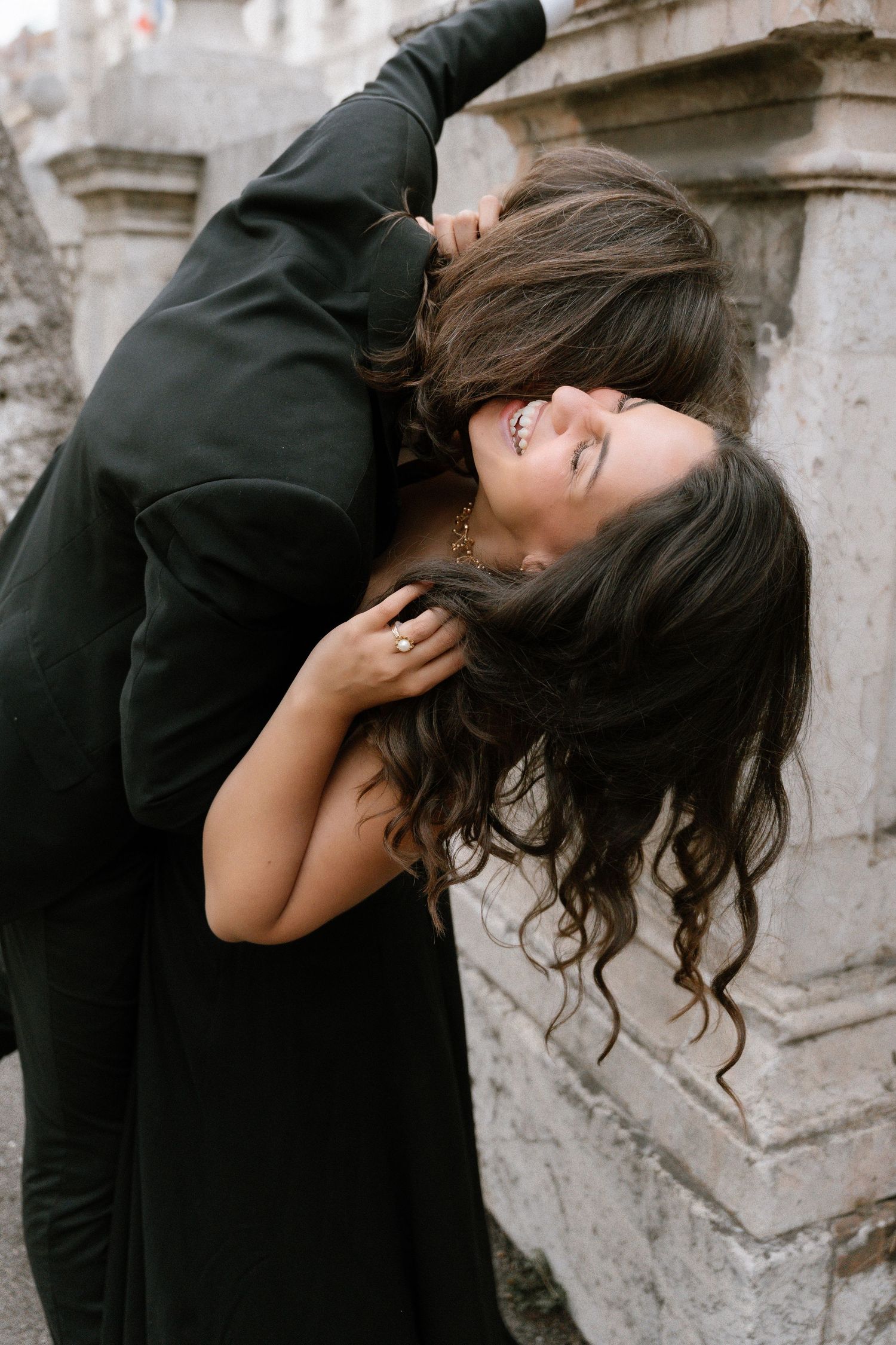 Two women in black embrace romantically near a stone wall, laughing joyfully in an outdoor setting.