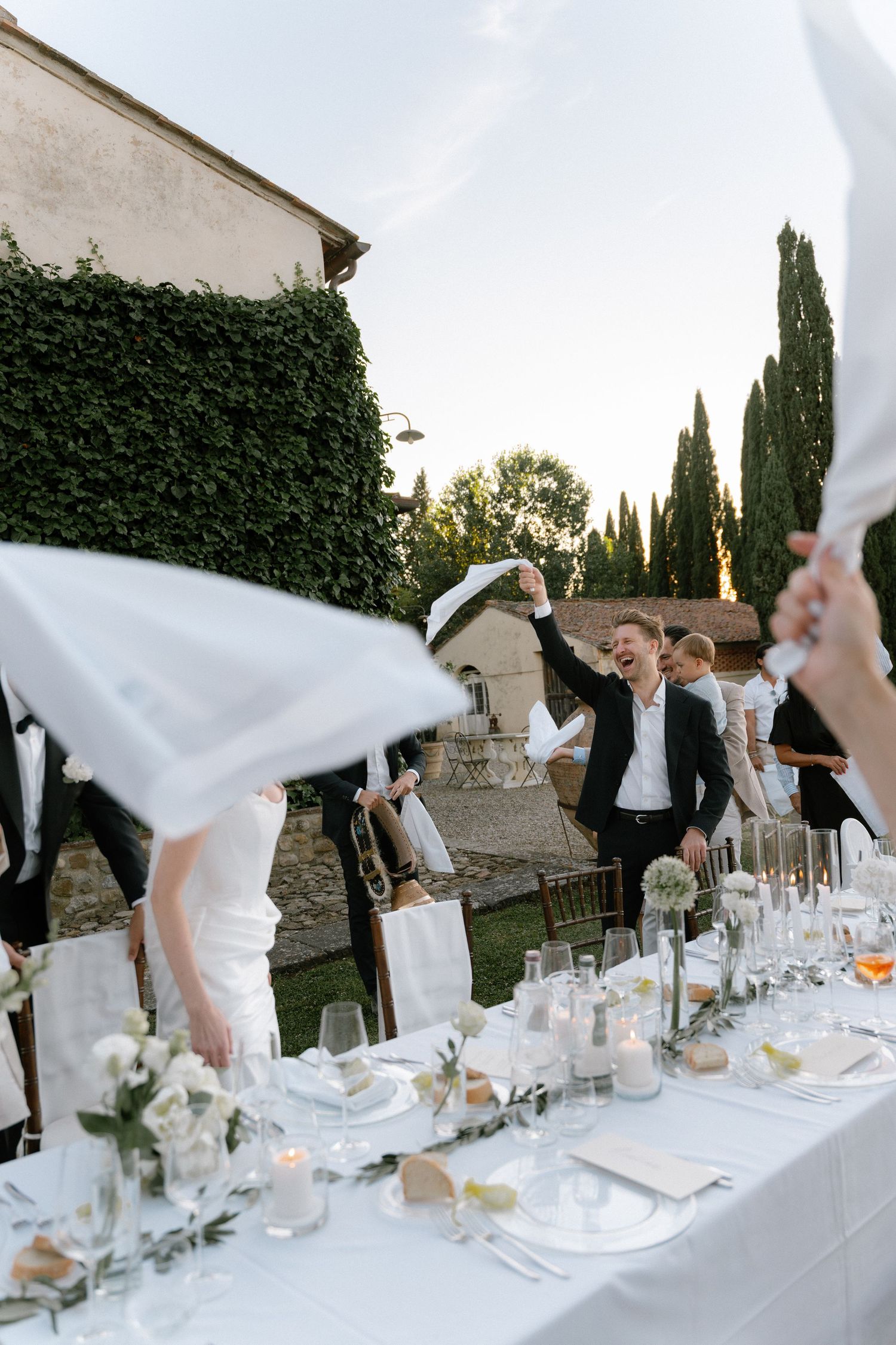 Elegant outdoor wedding reception with guests waving white napkins over a beautifully set table in a garden setting.
