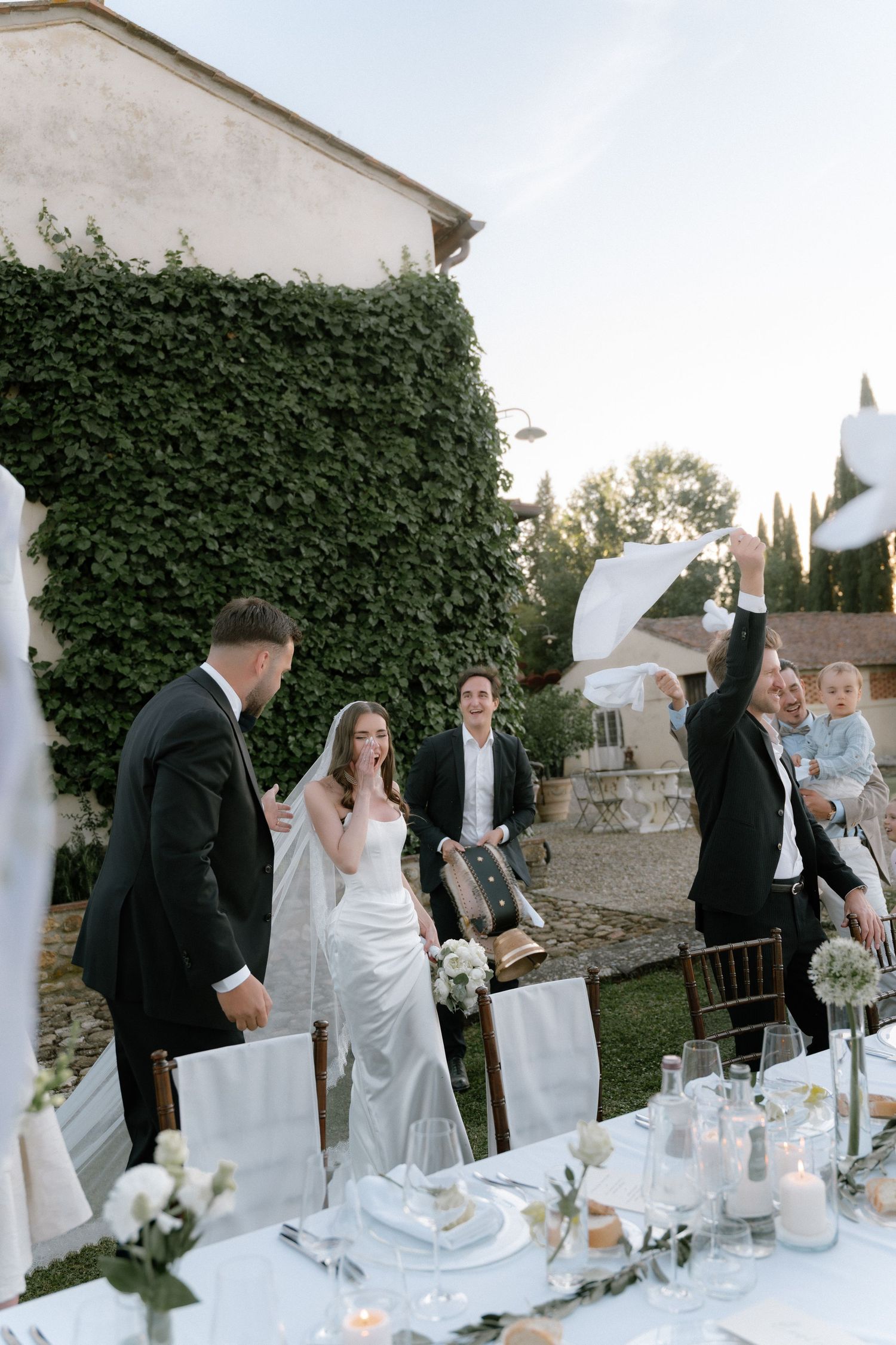 Outdoor wedding reception with bride and groom celebrating at elegantly set table, ivy-covered wall backdrop, Tuscany villa.