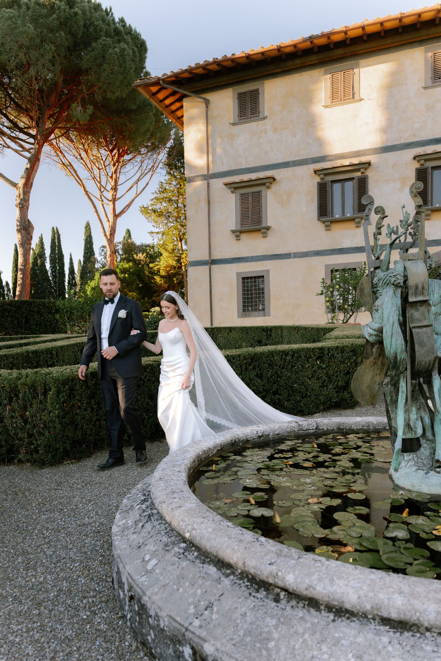 Bride and groom walk beside ornate fountain in lush Italian villa garden, bride wearing long veil and white gown.