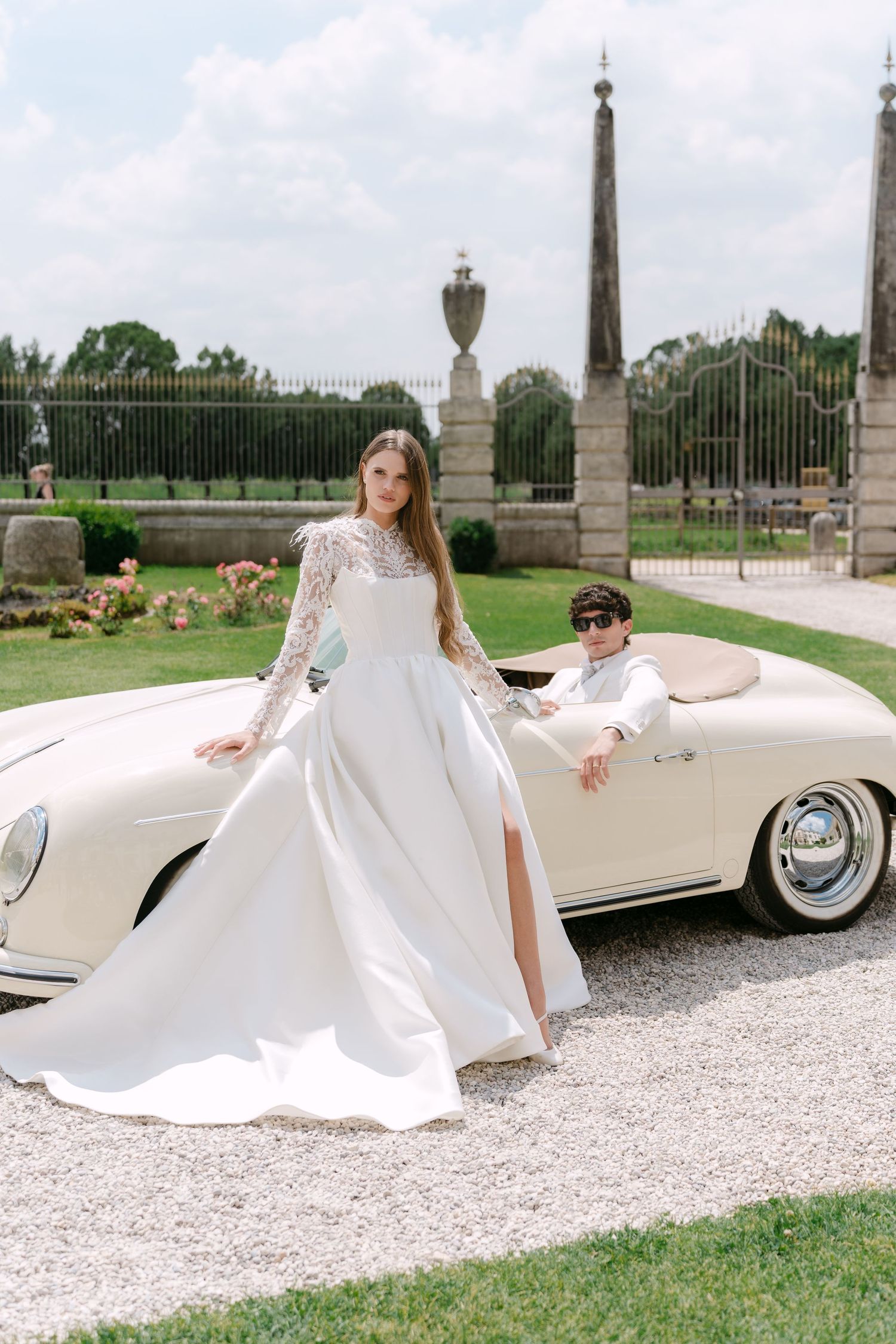 Elegant wedding scene with flowing white gown and classic convertible on gravel driveway.