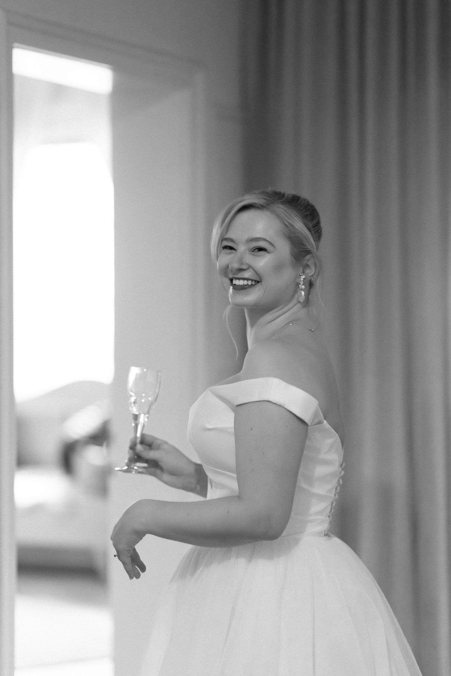 A joyful bride in a white wedding dress laughs while holding a champagne glass in a sunlit room.