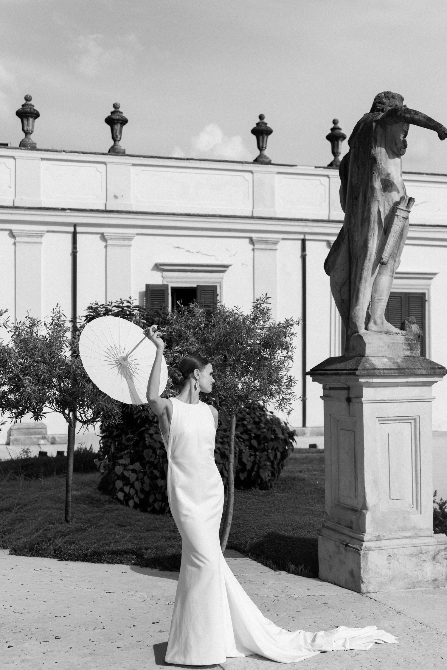 Elegant bride in white mermaid dress poses with parasol next to classical stone statue in garden setting. Villa Mosconi Bertani