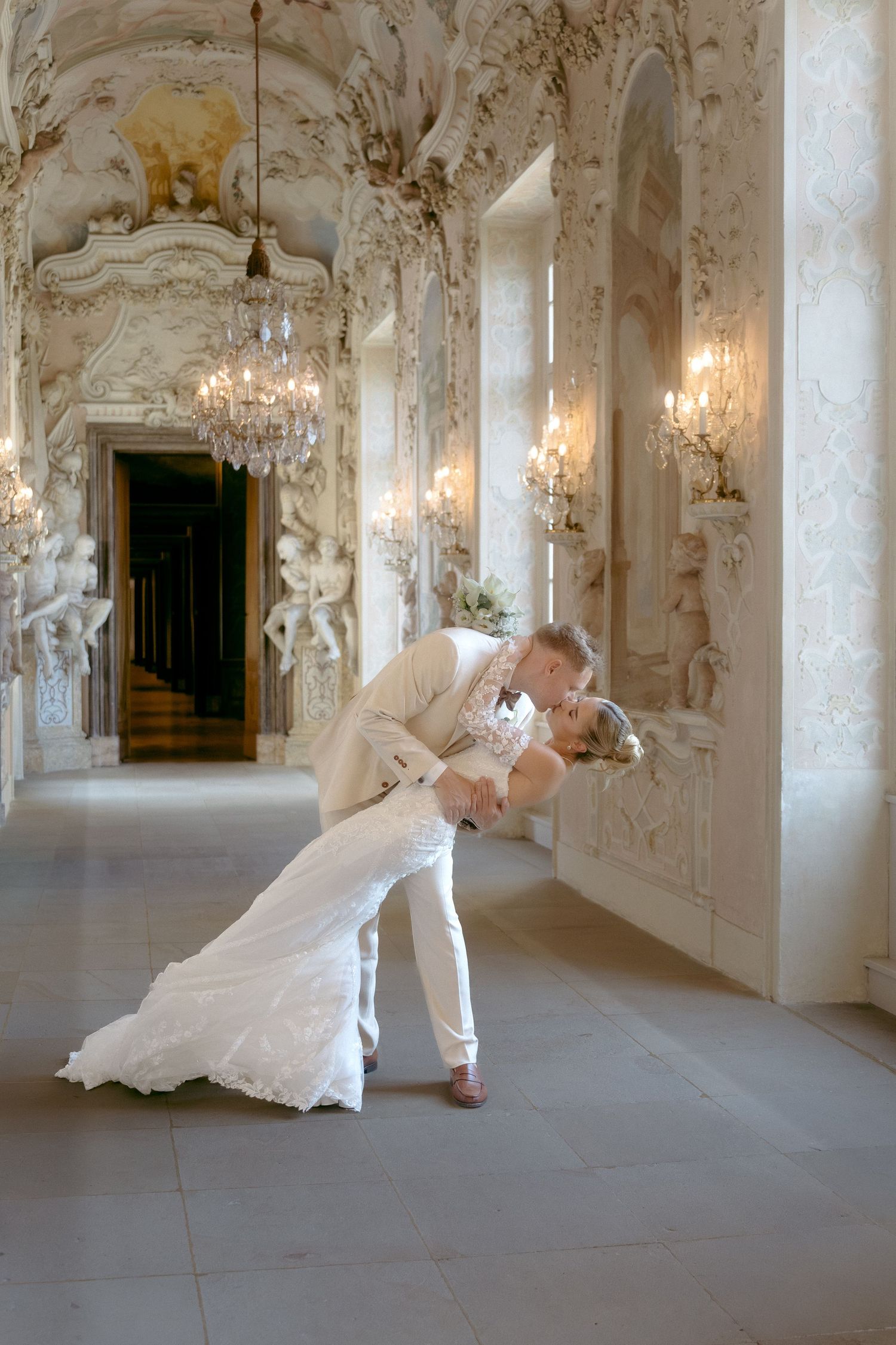 Elegant figures in flowing white gowns dance gracefully through an ornate baroque palace hallway with crystal chandeliers.Schloss Ludwigsbug