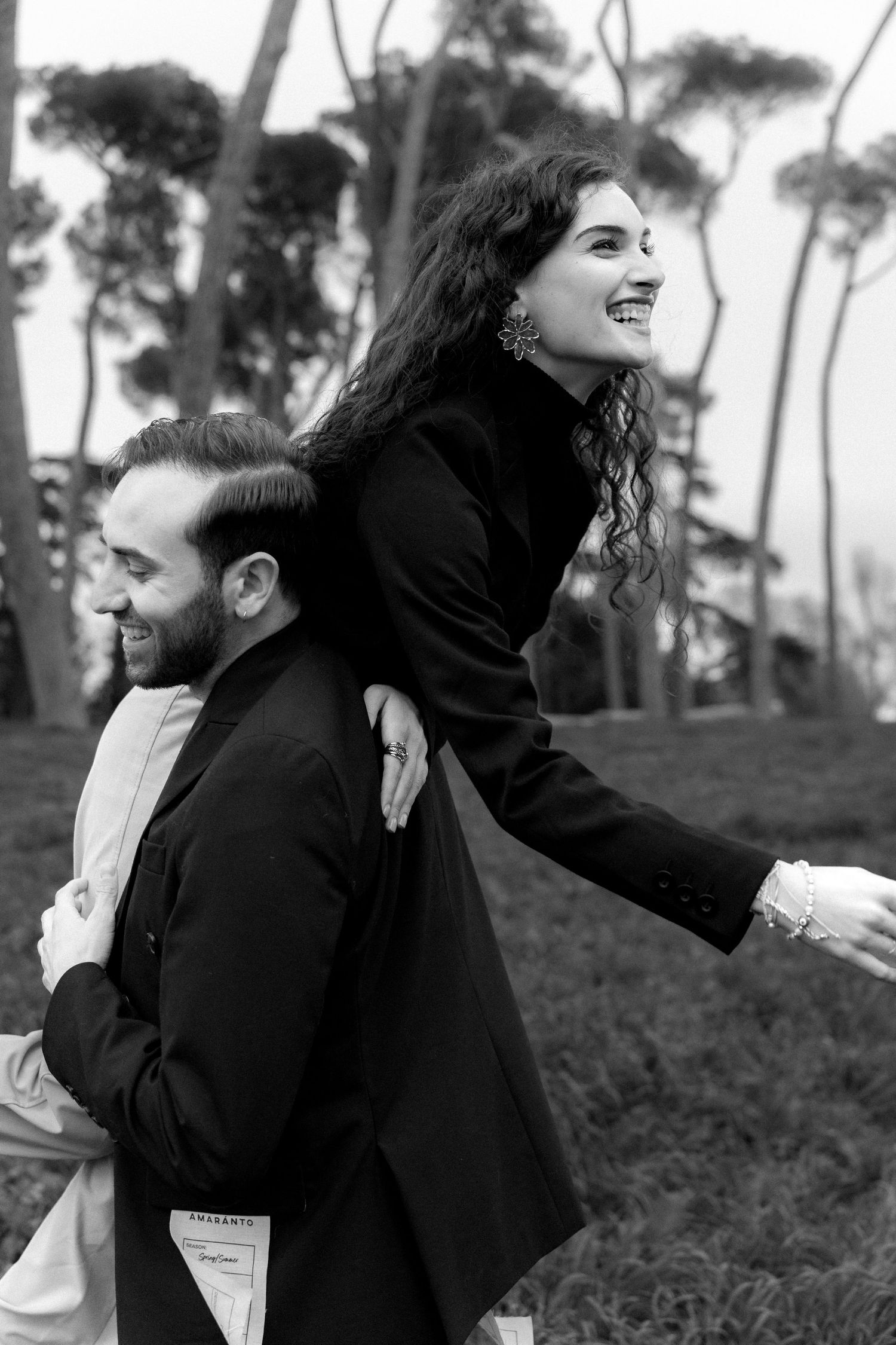 A joyful couple in black coats shares a playful piggyback ride moment outdoors with trees in the background.