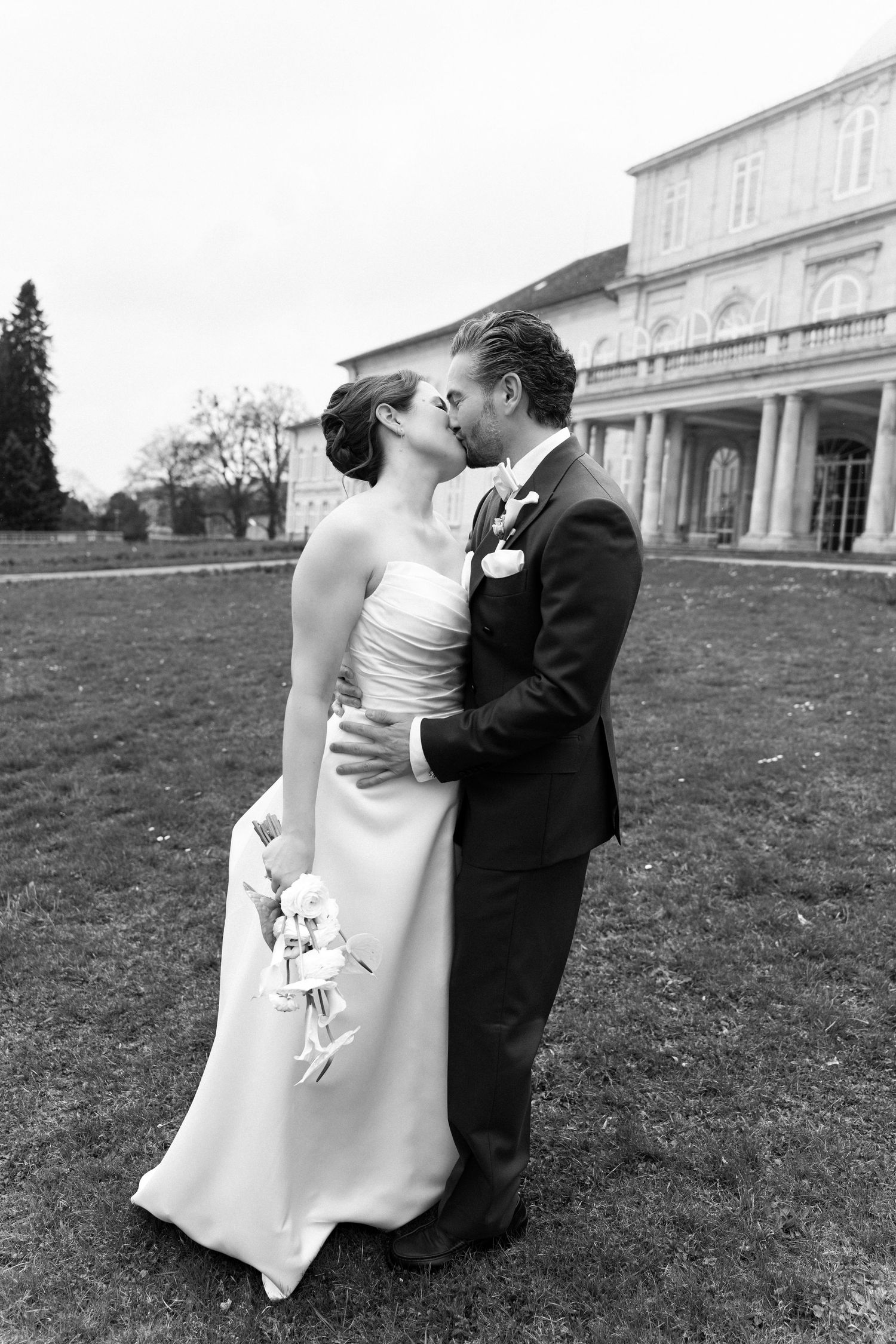 Black and white photo of a bride and groom sharing a kiss on a lawn outside an elegant building on their wedding day.