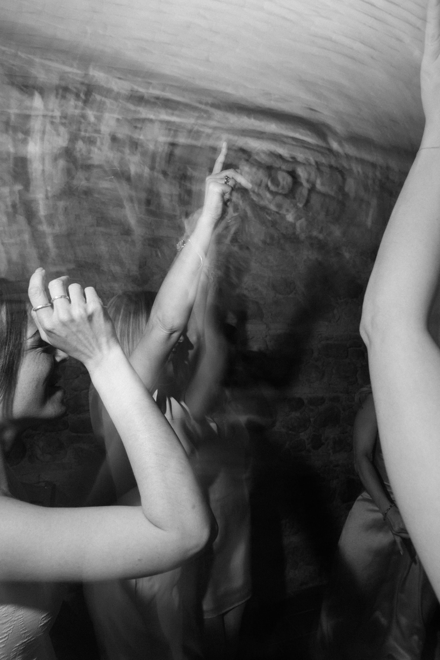 Black and white blurred photo of people dancing with raised arms at a concert or party event.