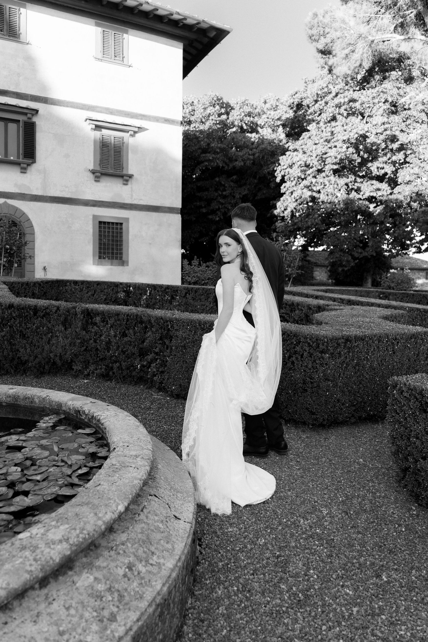 Black and white photo of bride and groom in formal wear strolling through elegant Italian villa garden with manicured hedges.