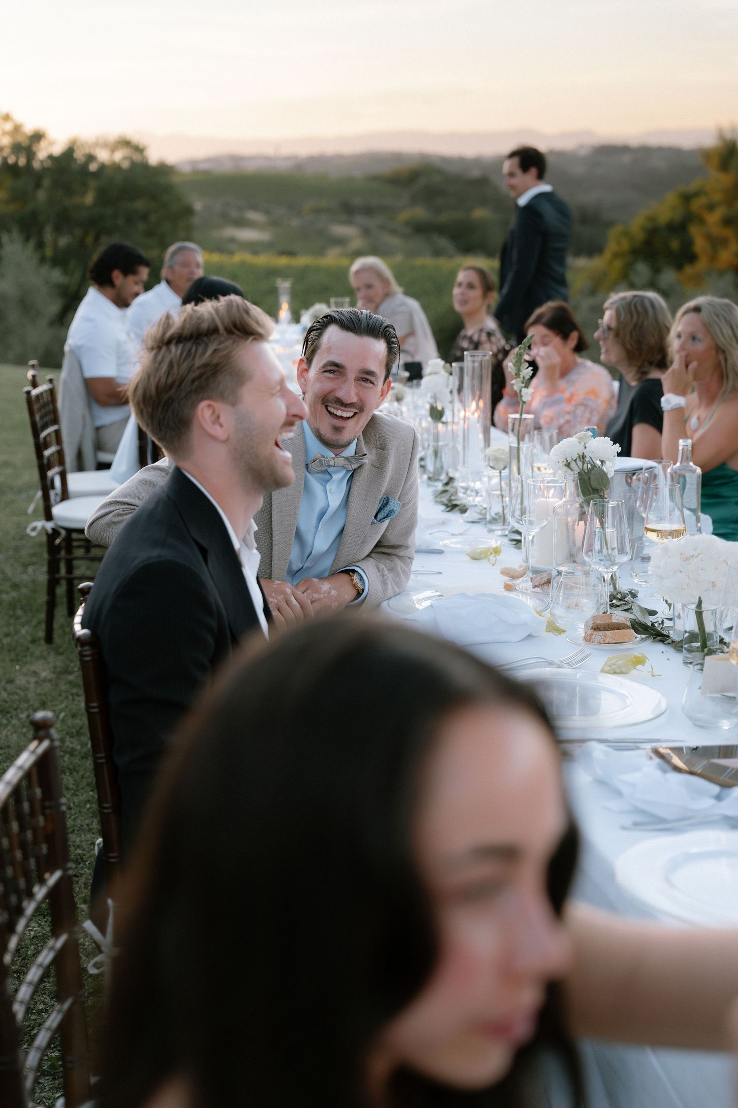Two men laughing joyfully at an elegant outdoor wedding reception dinner table at sunset.