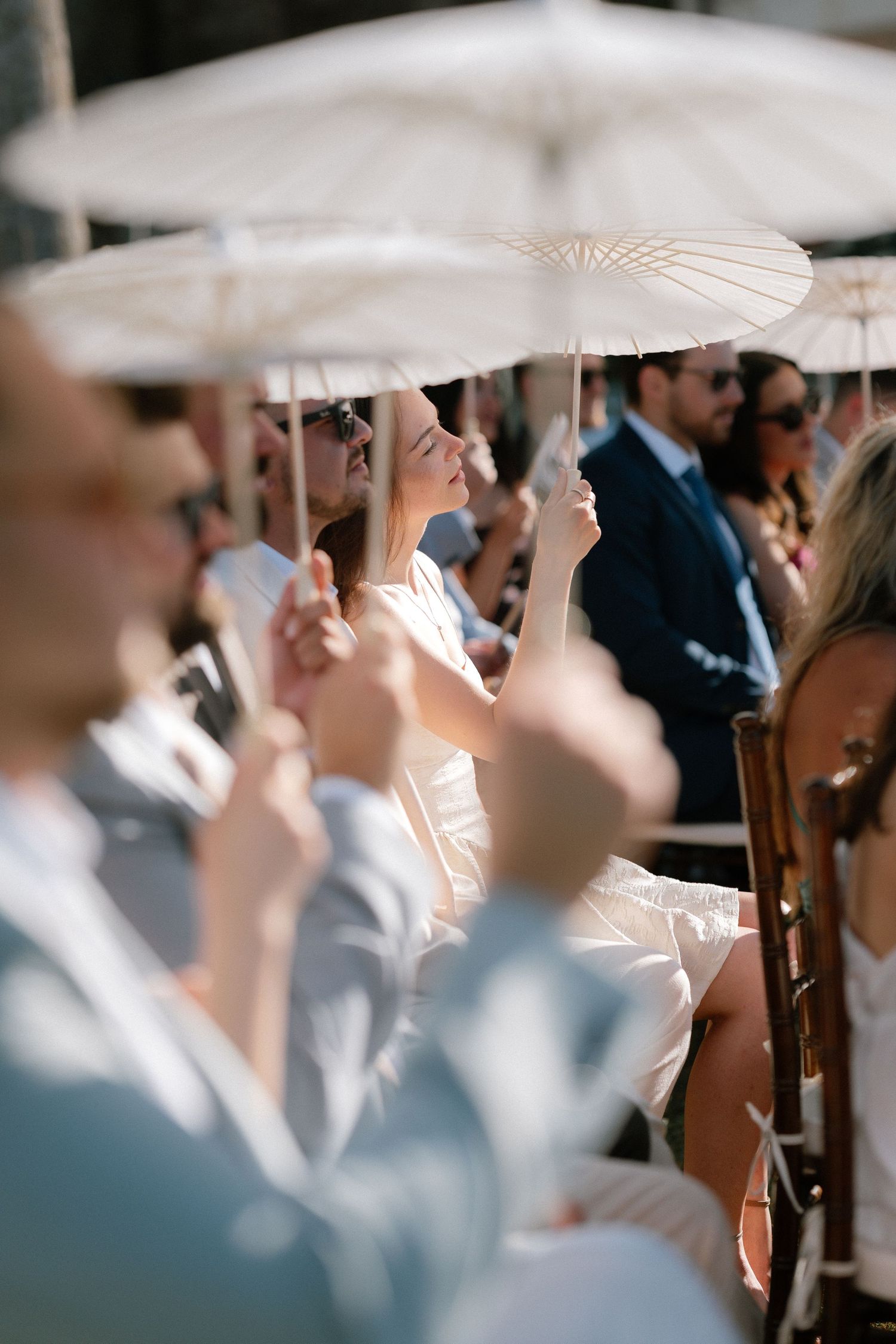 Wedding guests holding white parasols while seated outdoors in the sun during a ceremony.