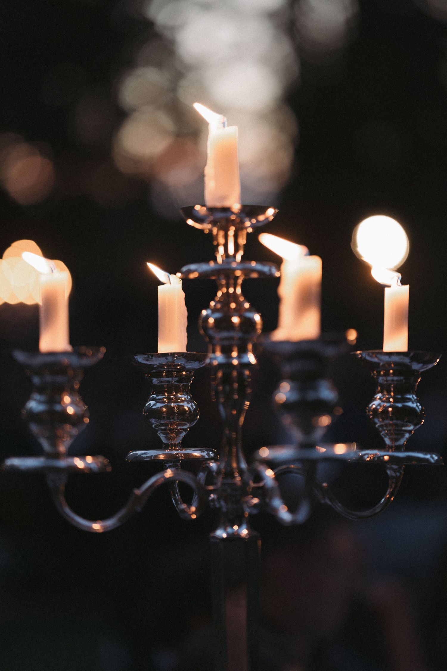 Silver candelabra with lit white candles glowing softly against a dark background with warm bokeh lights.
