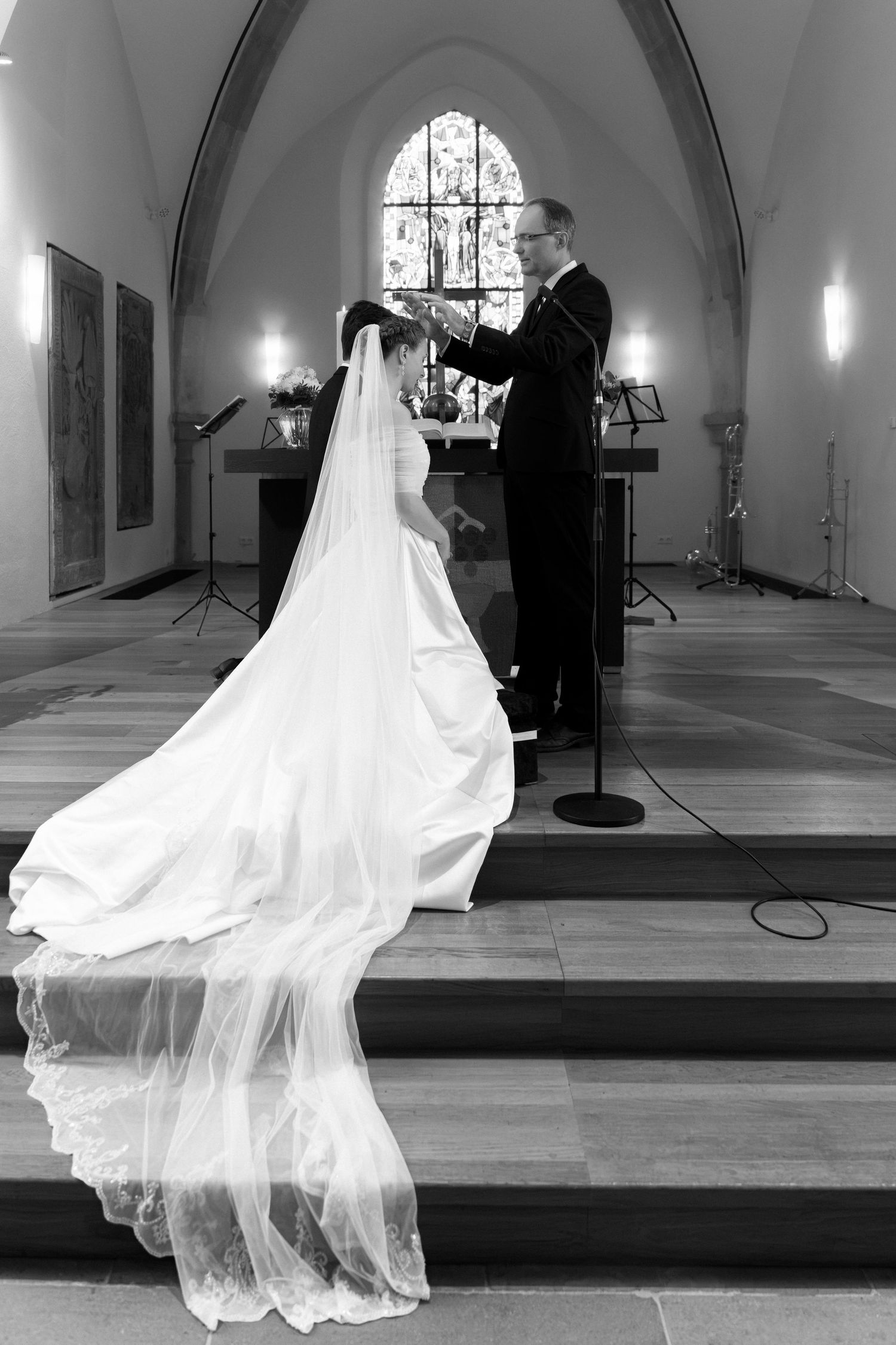 Black and white wedding photo of a bride in a long lace veil kneeling at a church altar as a pastor blesses her.