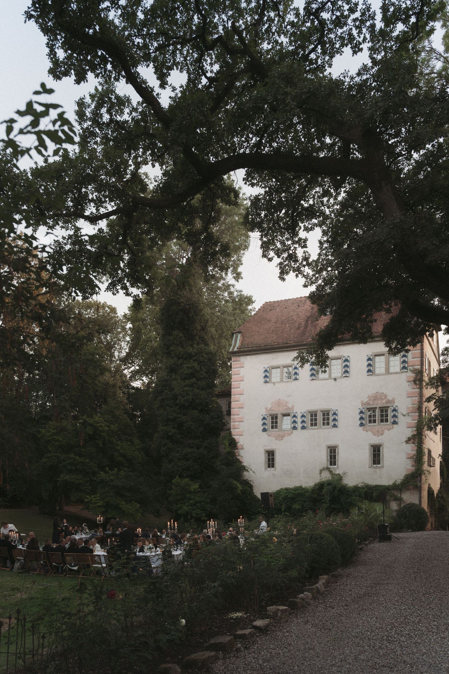 Guests gather for an outdoor dinner under tall trees beside a historic white castle with shuttered windows and a gravel path at dusk.