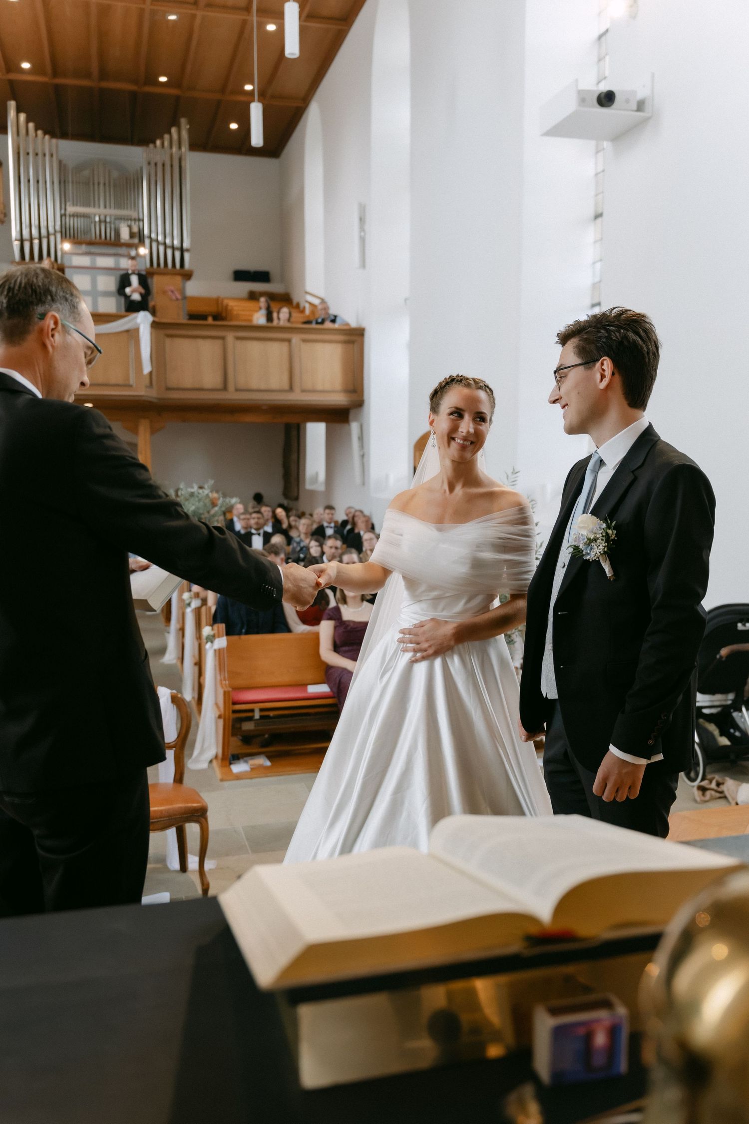 Smiling bride in an off-shoulder white wedding gown shakes hands with an officiant beside the groom during a church ceremony.