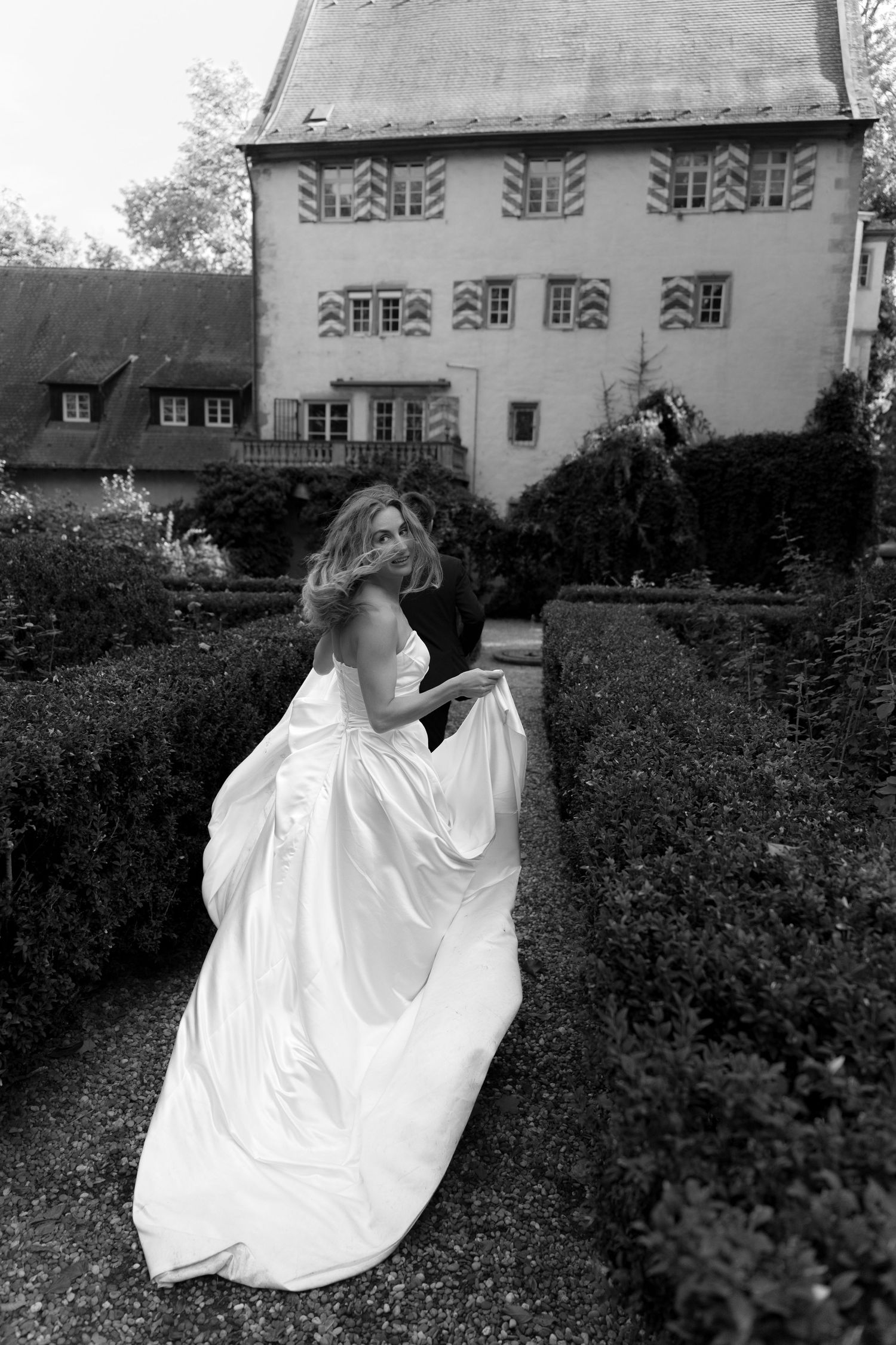 Black and white photo of a bride in a flowing satin wedding gown walking through a hedge garden toward a historic castle-style building.
