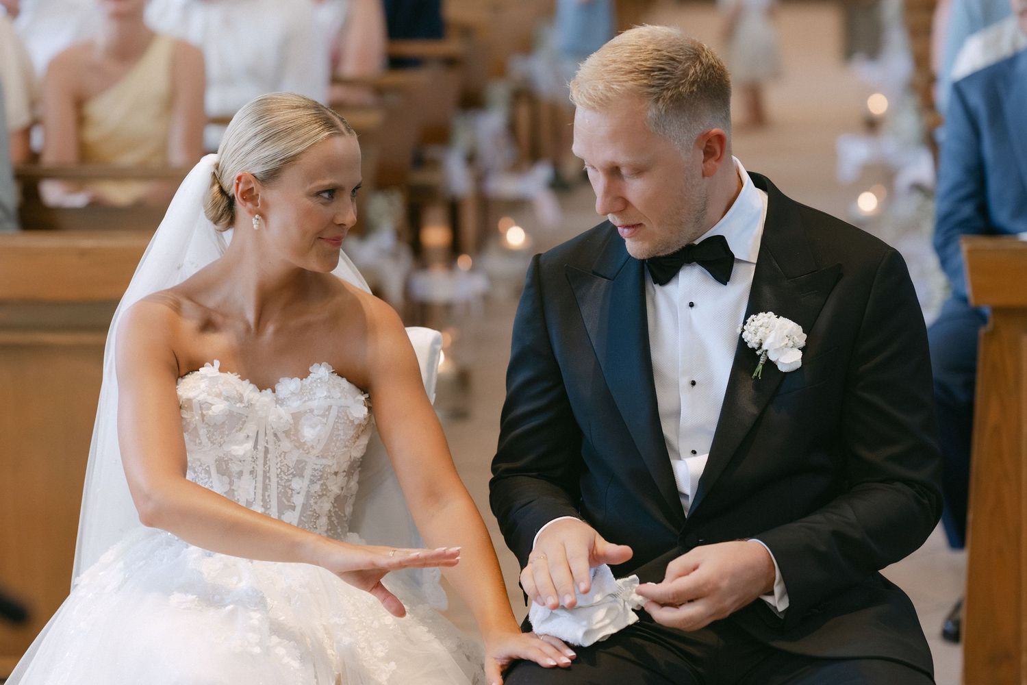 Bride in a strapless lace wedding gown and groom in a black tuxedo with bow tie sit in a church pew during their wedding ceremony.