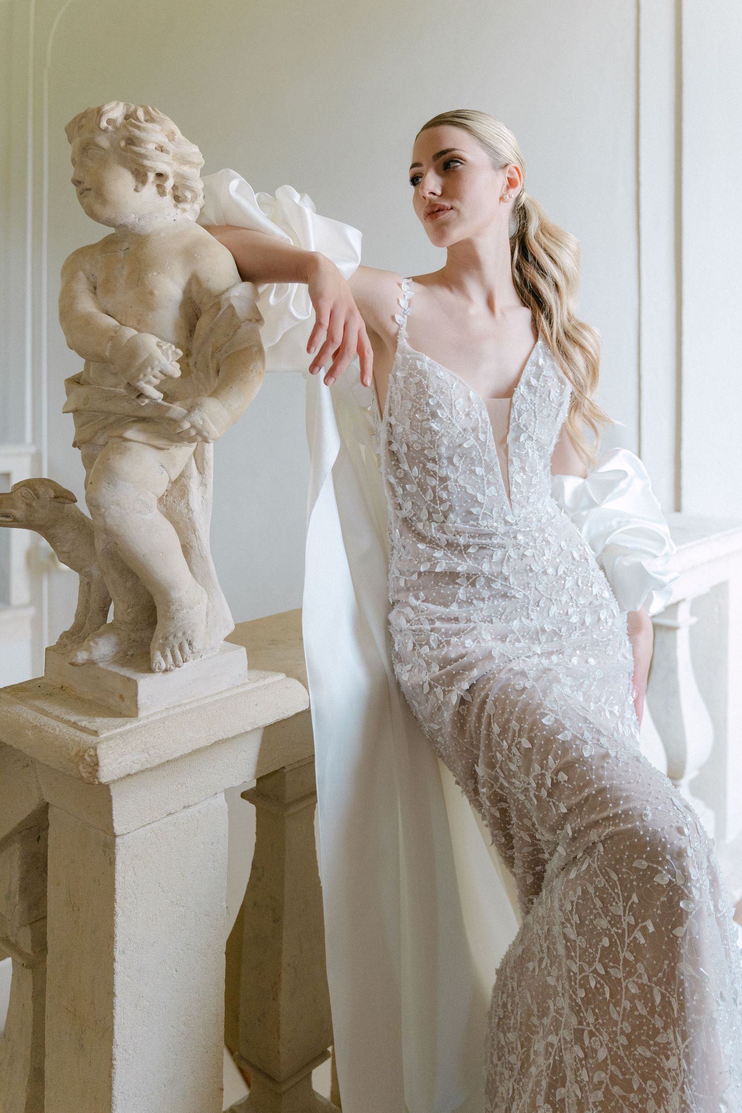 Elegant bridal fashion shoot featuring lace gown next to classical stone sculptures.