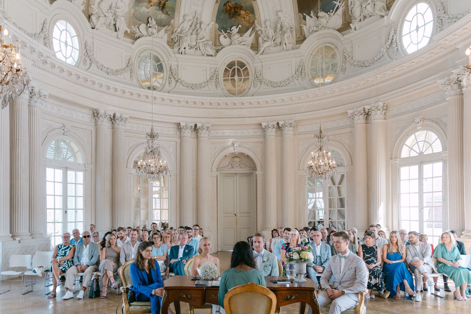 Panoramic view of an ornate white ballroom with crystal chandeliers and guests gathered for an elegant event. Schloss Solitude