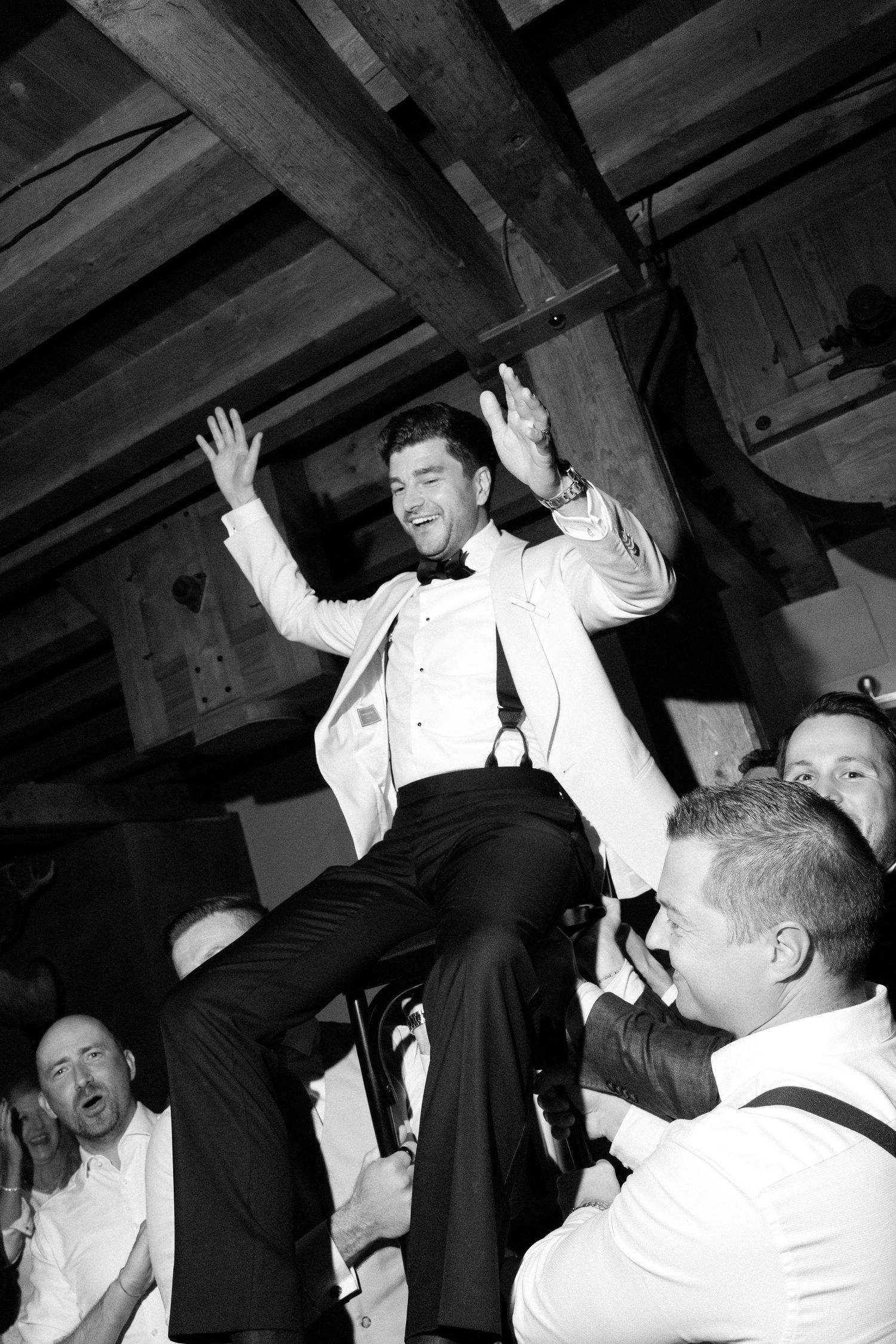 A joyful celebration at a wedding reception shows a guest being lifted up by others during a festive dance moment.