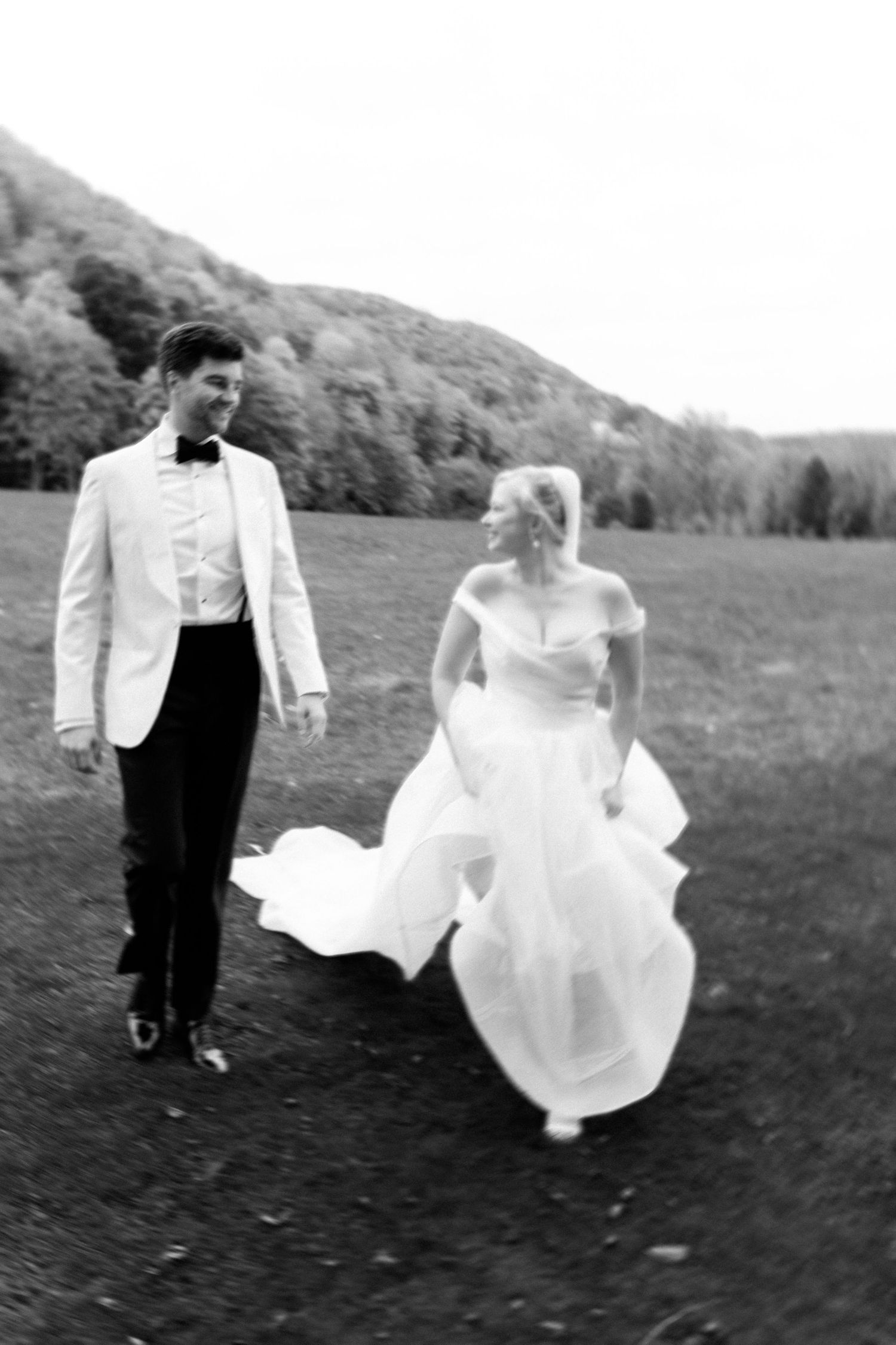 A bride in a flowing white dress and groom in black tuxedo walk together across a grassy hillside in a series of candid shots.