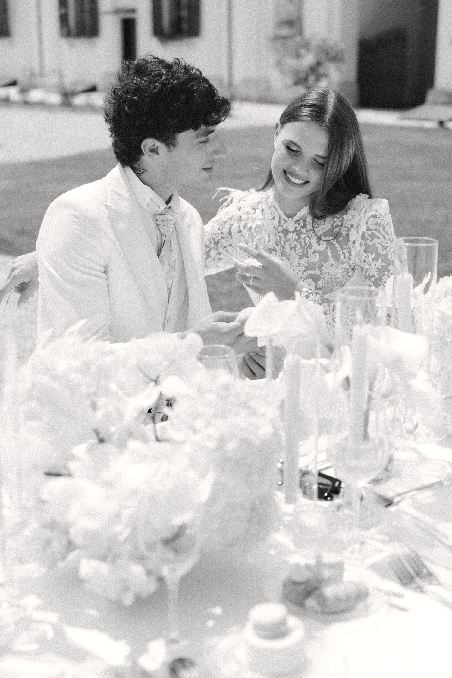 A couple shares a joyful moment at an elegant outdoor table setting with white floral arrangements. Villa Mosconi Bertani
