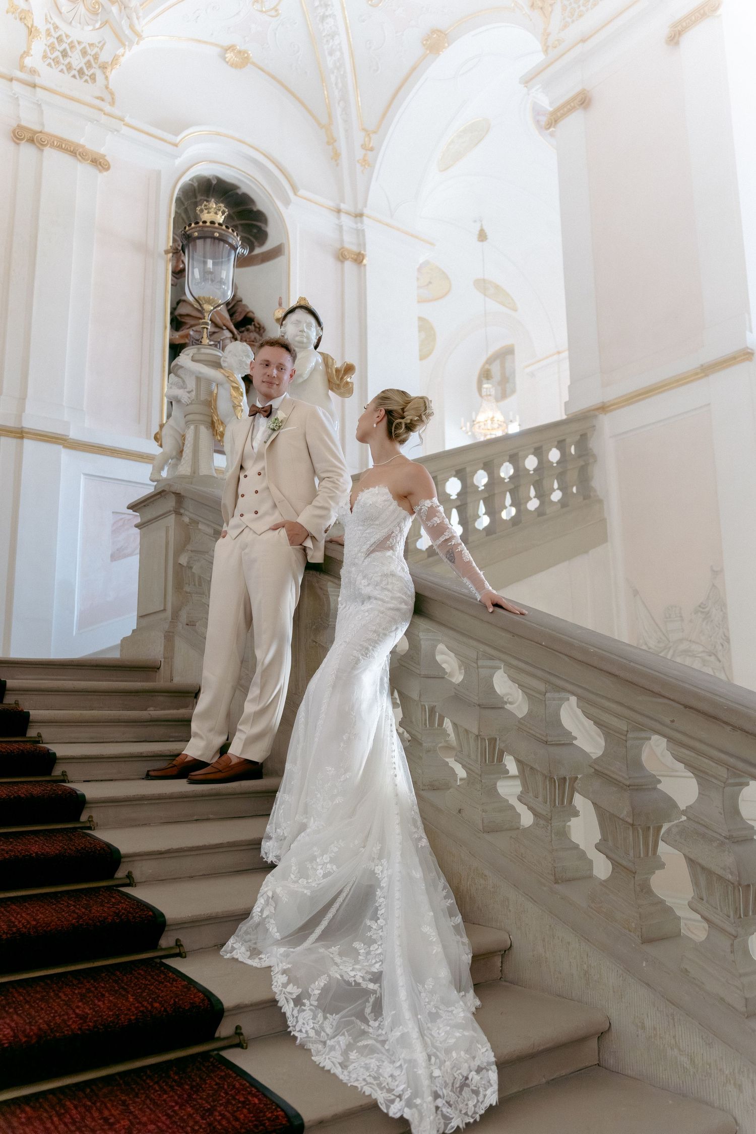 A bride in a flowing lace wedding gown gracefully ascends an elegant white marble staircase in a historic building. Schloss Ludwigsburg