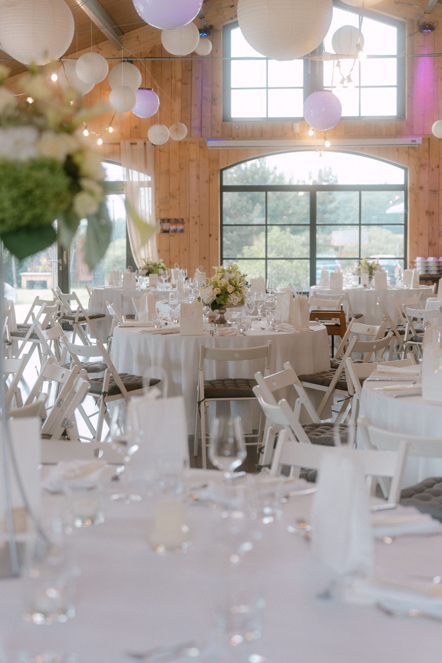 Elegant wedding reception venue with white tables, paper lanterns, and natural light streaming through large windows.