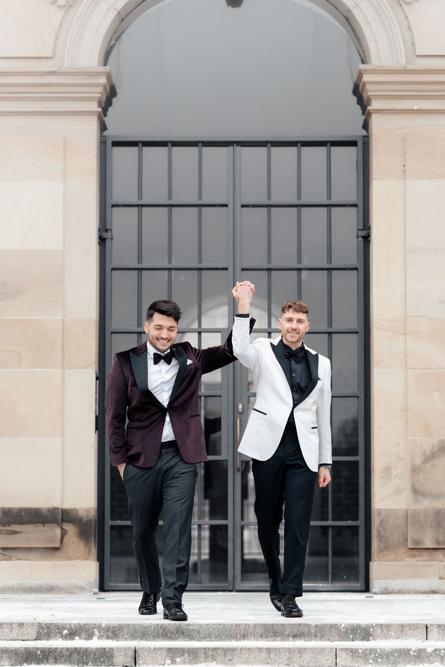 Two people in formal tuxedos and bow ties celebrate on stone steps in front of an arched metal gate.