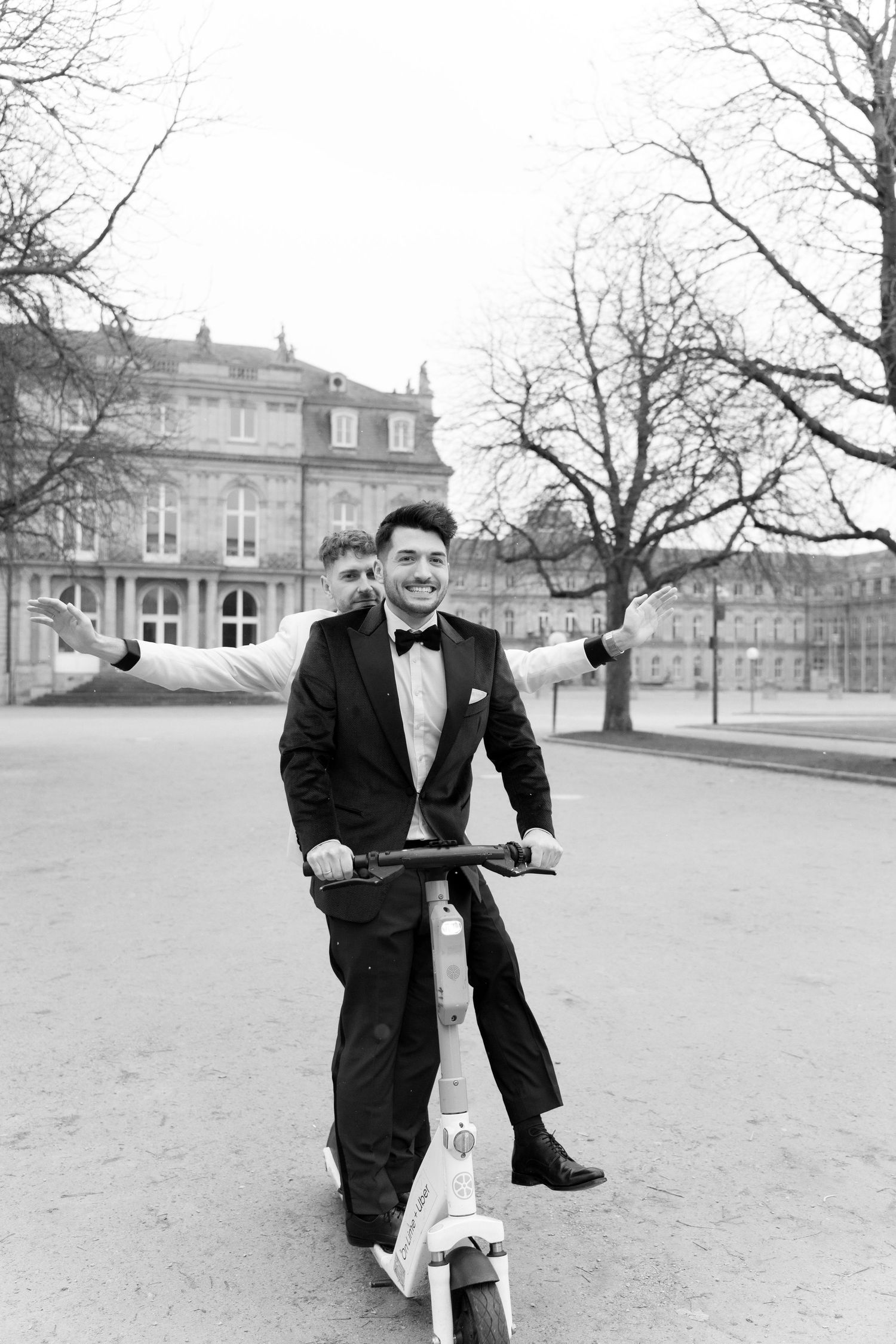 A person in a tuxedo smiles while riding a scooter in front of a grand historic building on a winter day.