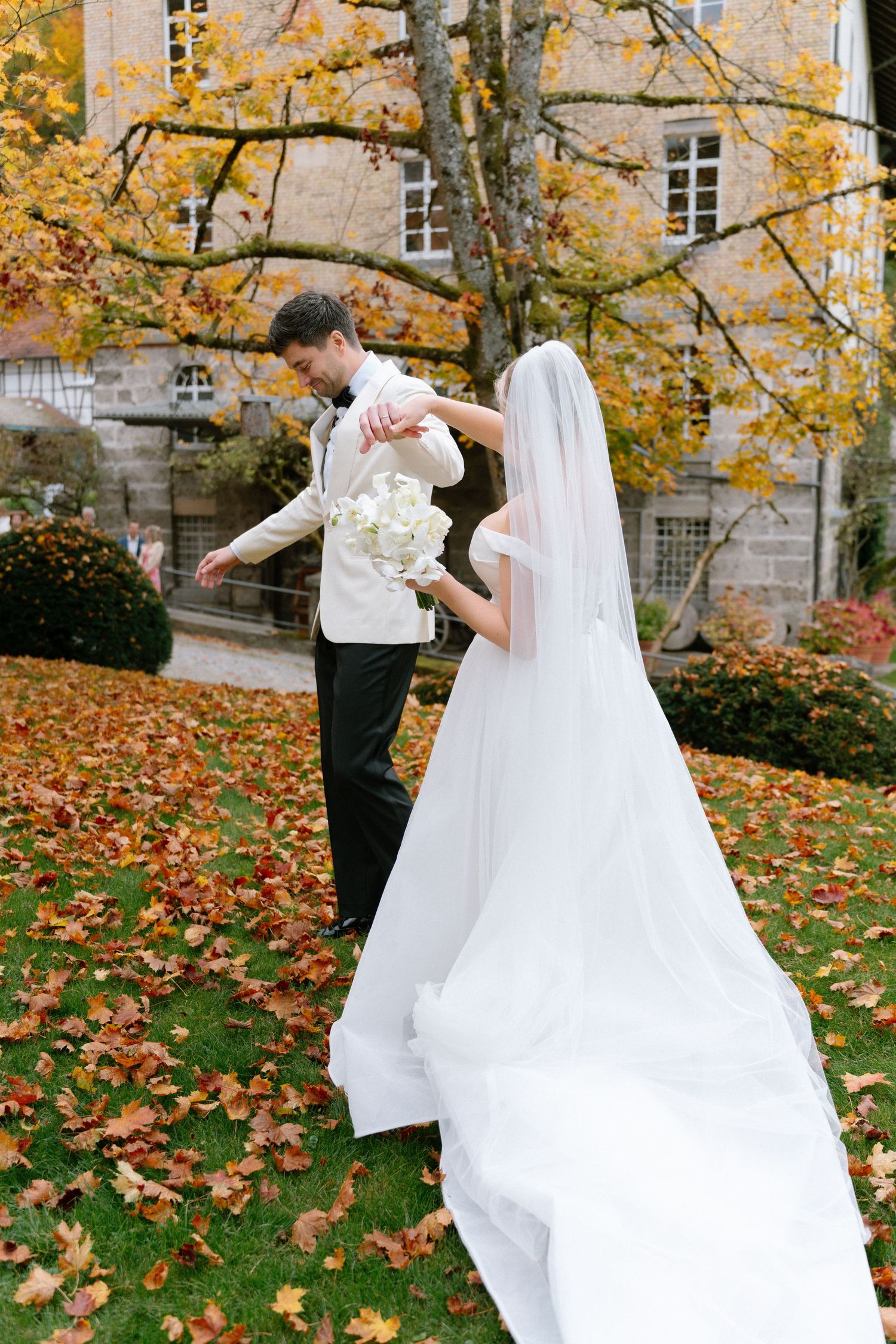 A couple in wedding attire dances together on fallen autumn leaves with a stone building in the background.