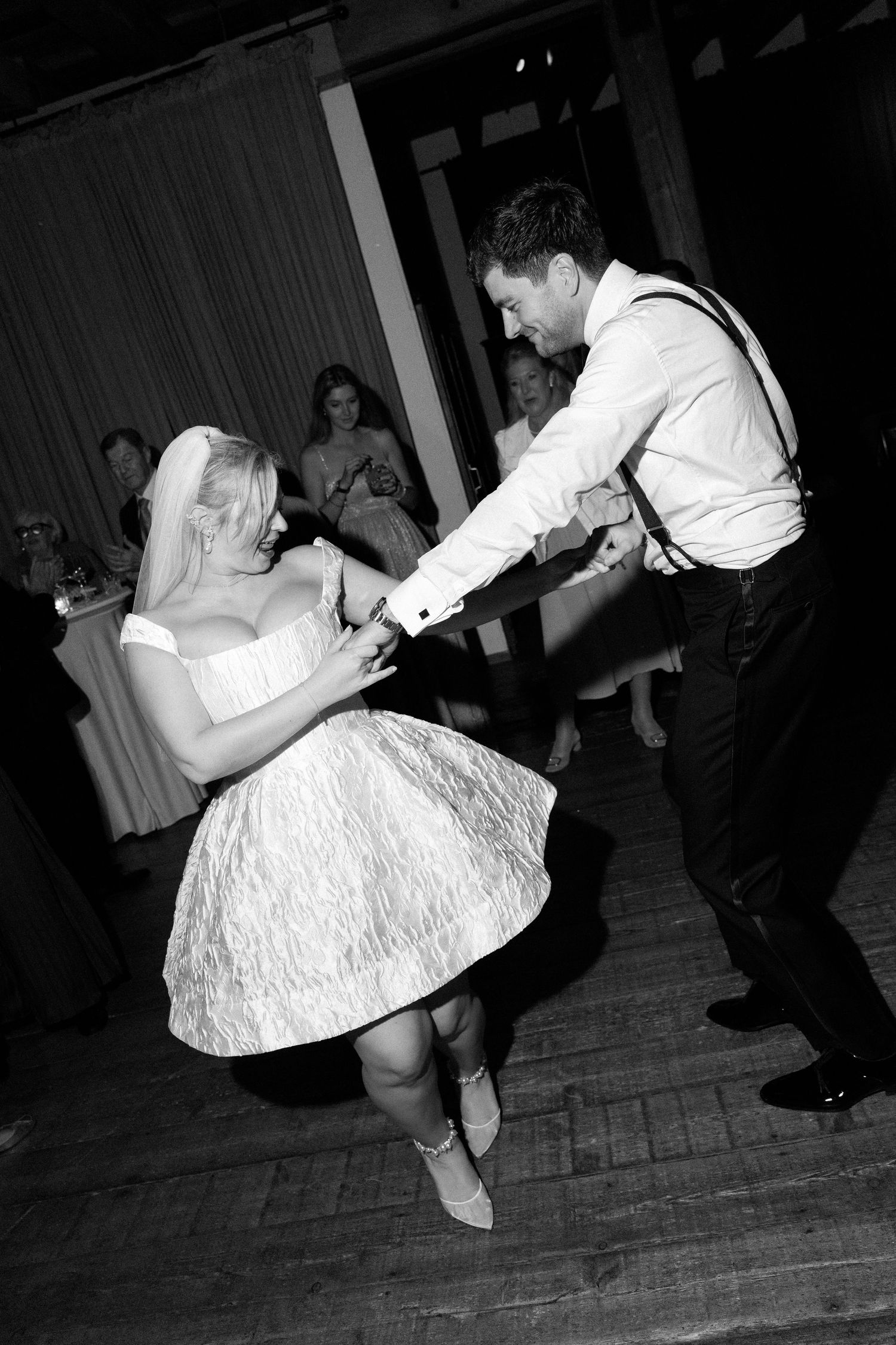 A couple dances energetically at a wedding reception, with the bride wearing a short white lace dress on a wooden dance floor.
