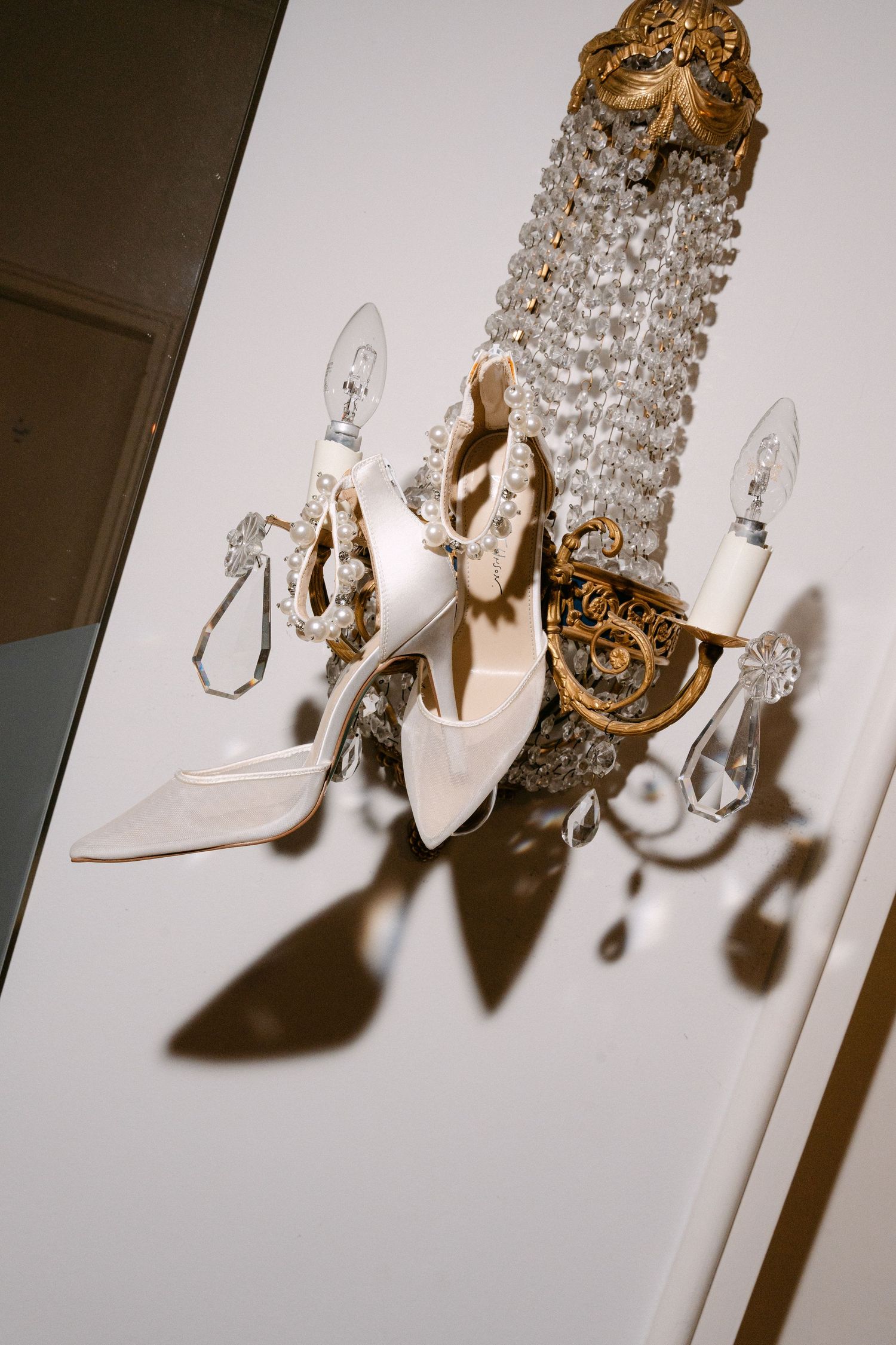 Elegant white bridal shoes displayed beneath a crystal chandelier in a romantic setting.