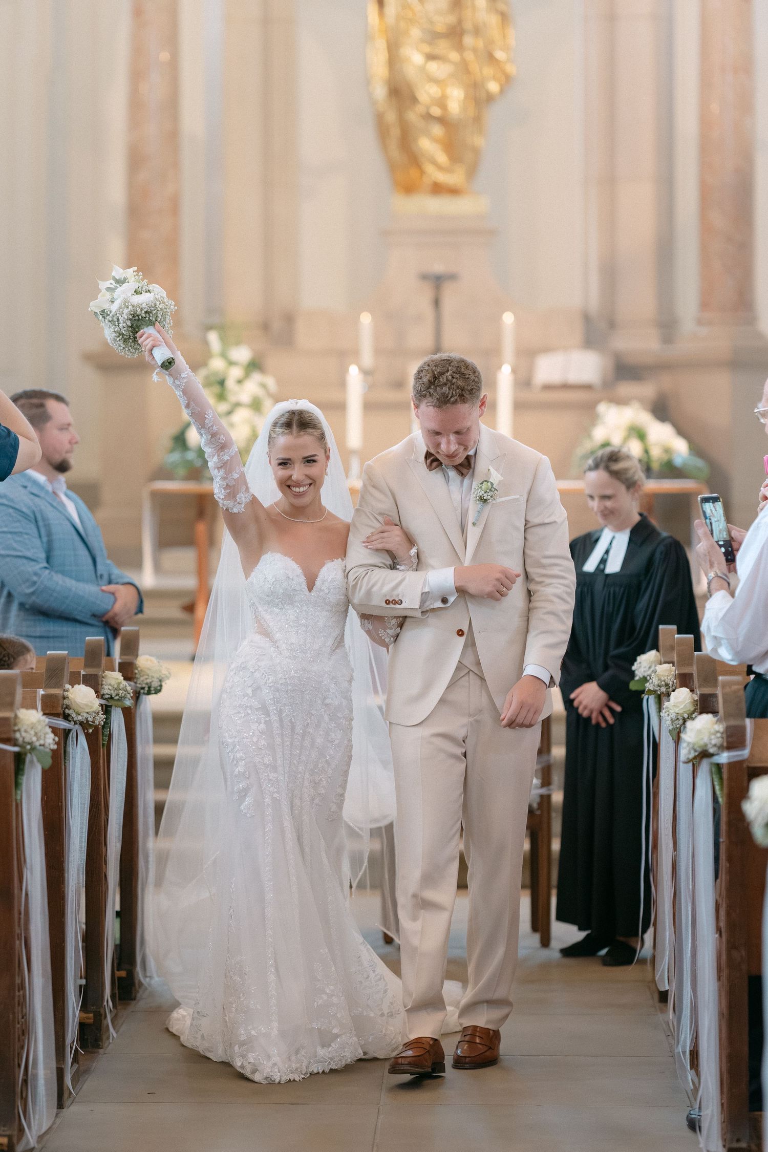 Wedding party members in white dresses and beige suits celebrate joyfully in a church aisle after ceremony.