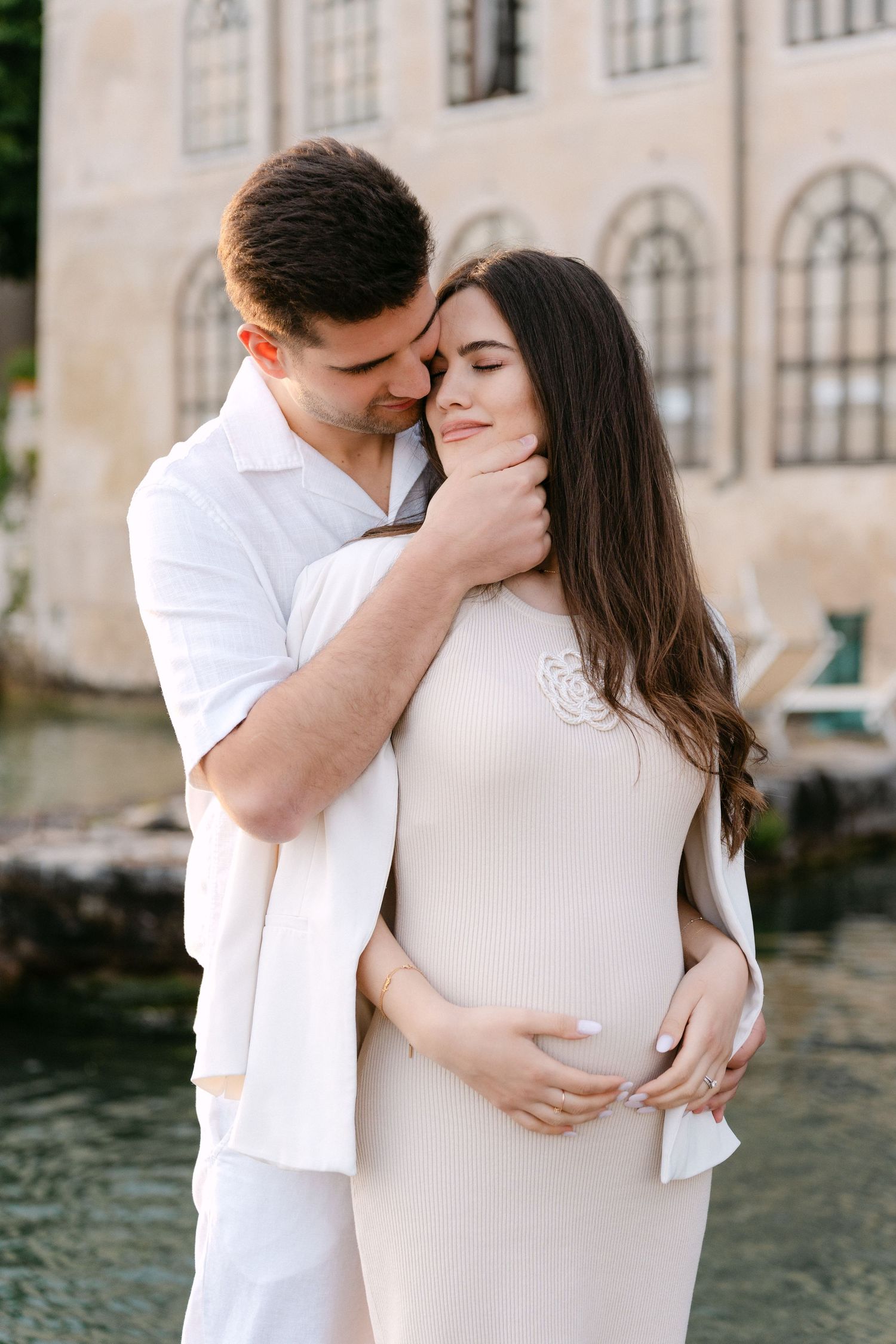 A romantic couple in white embraces and shares tender kisses by a waterfront building.