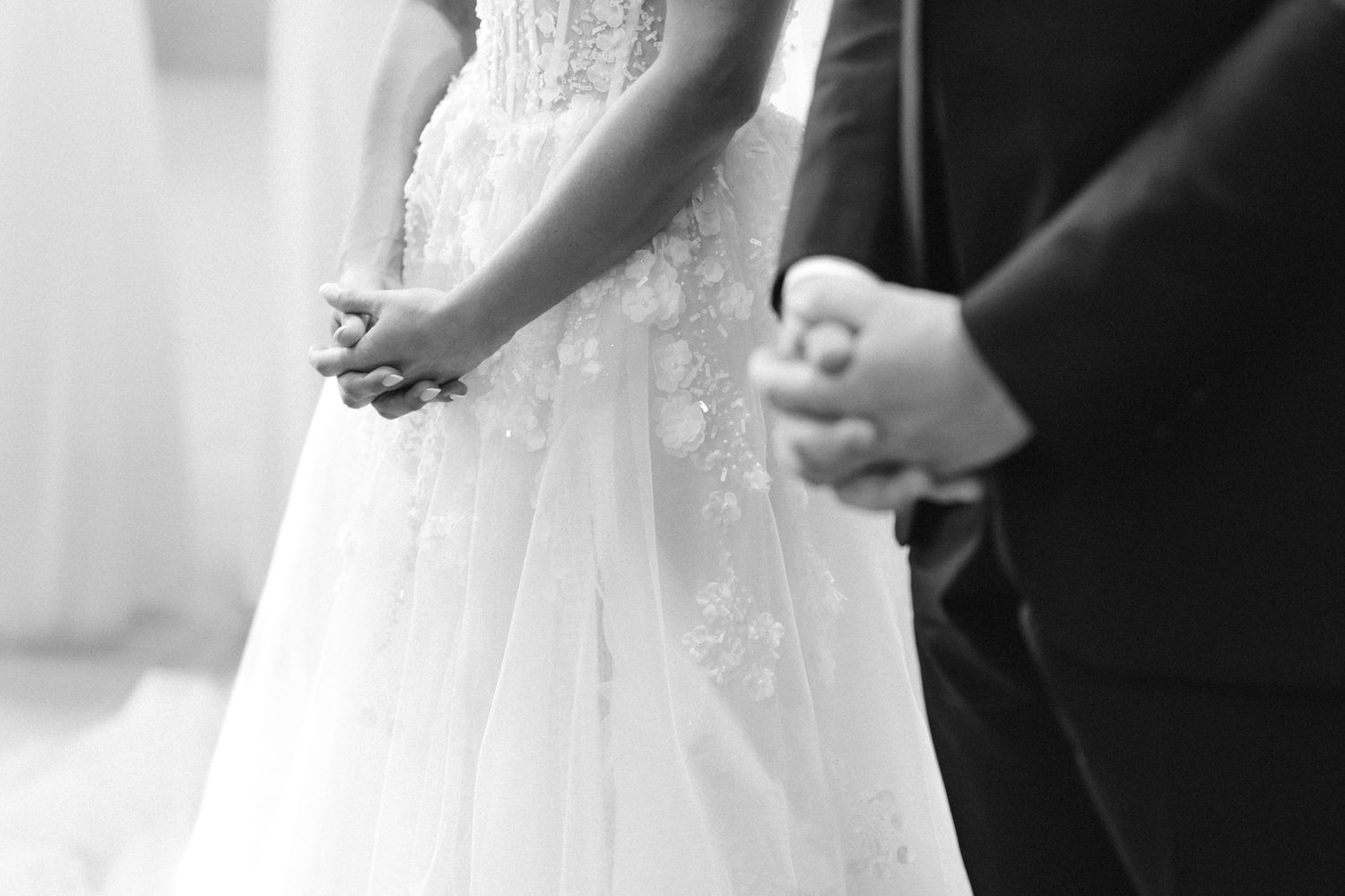 Black and white photo of a bride in a lace wedding gown and a groom in a dark suit standing close with hands clasped during a ceremony.