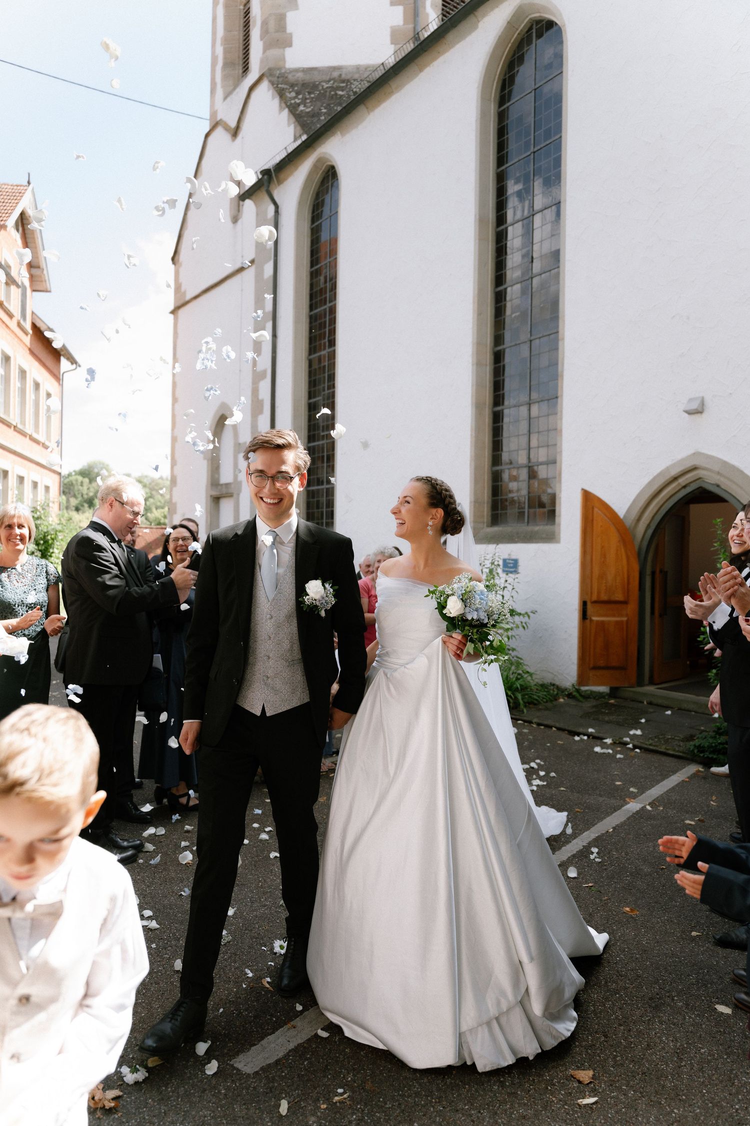 Bride in a white gown and groom in a dark suit walk past cheering guests tossing confetti outside a white stone church.