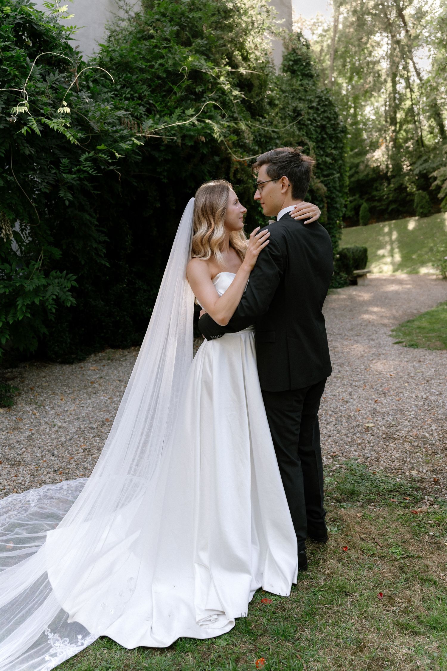 Bride in a white satin gown with a long veil embraces her groom in a black suit on a garden gravel path surrounded by greenery.